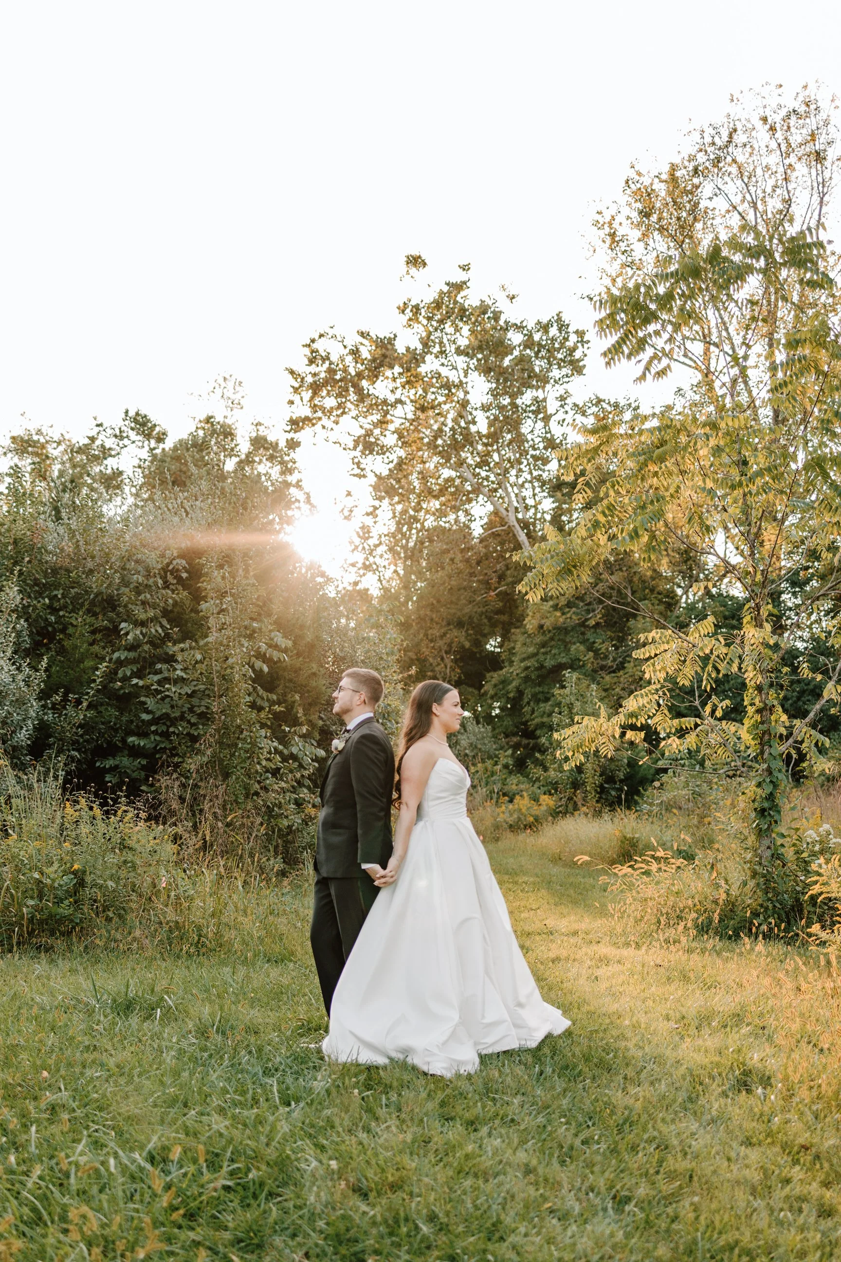 A bride and groom holding hands and standing back-to-back in a grassy outdoor setting during sunset, surrounded by trees.