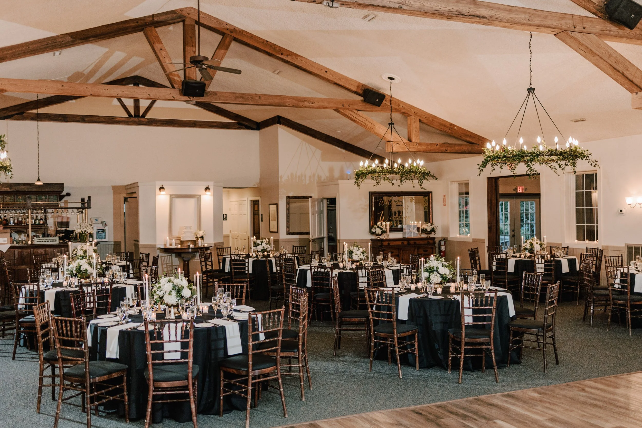 Wedding reception hall decorated with black tablecloths, white accents, floral centerpieces, and candle candles. Wooden chairs surround each table, and chandeliers with greenery hang from the ceiling. The room has large windows and a bar area on the 