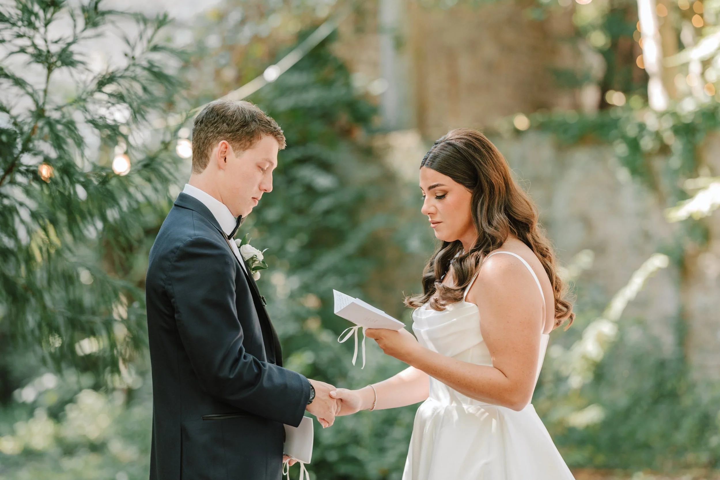 A bride and groom exchanging vows outdoors with greenery in the background.