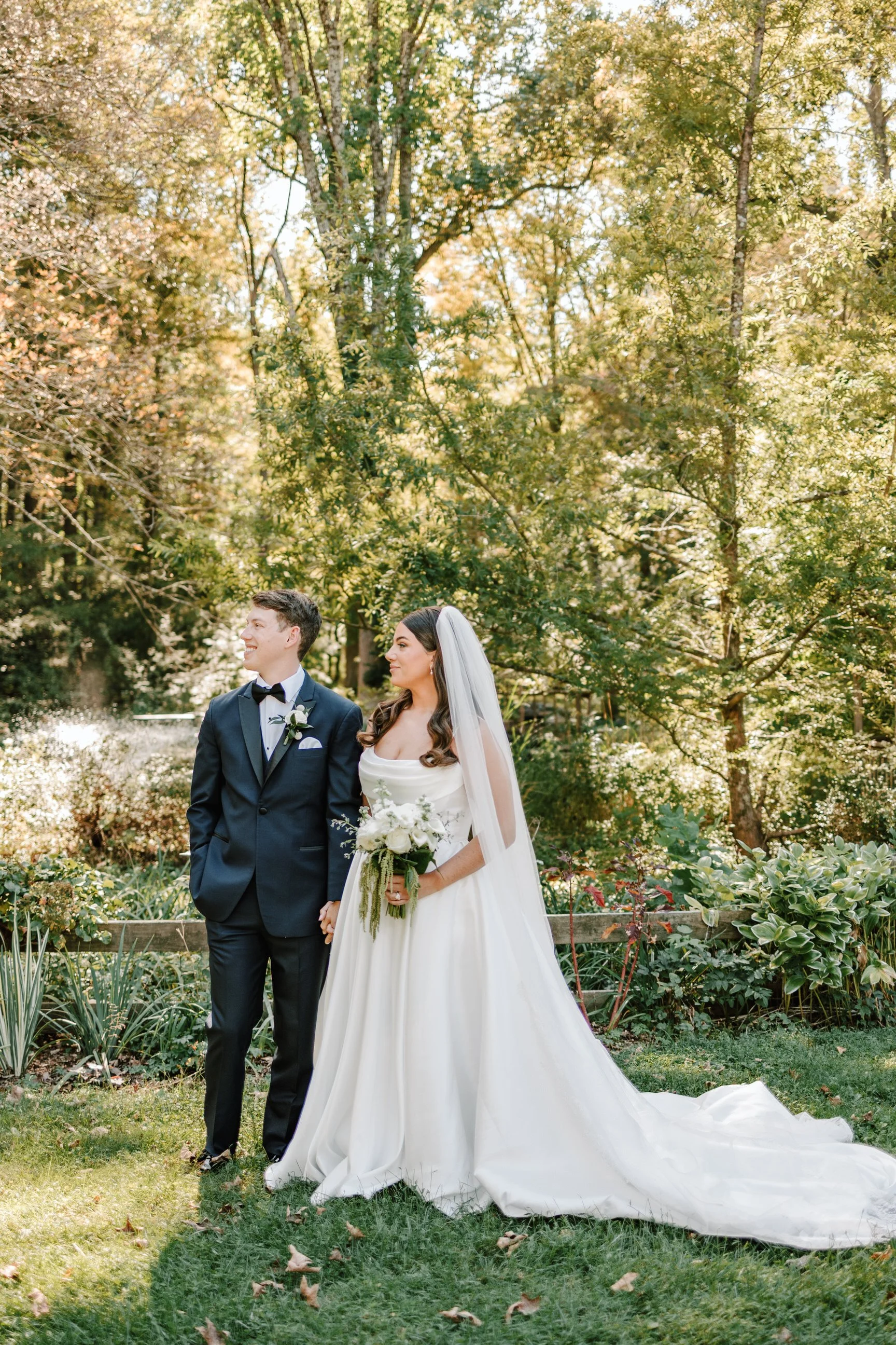 A bride and groom standing outdoors in a garden during daytime, holding hands. The bride is in a white wedding gown with a long train and veil, holding a bouquet of white flowers. The groom is in a black tuxedo, smiling.