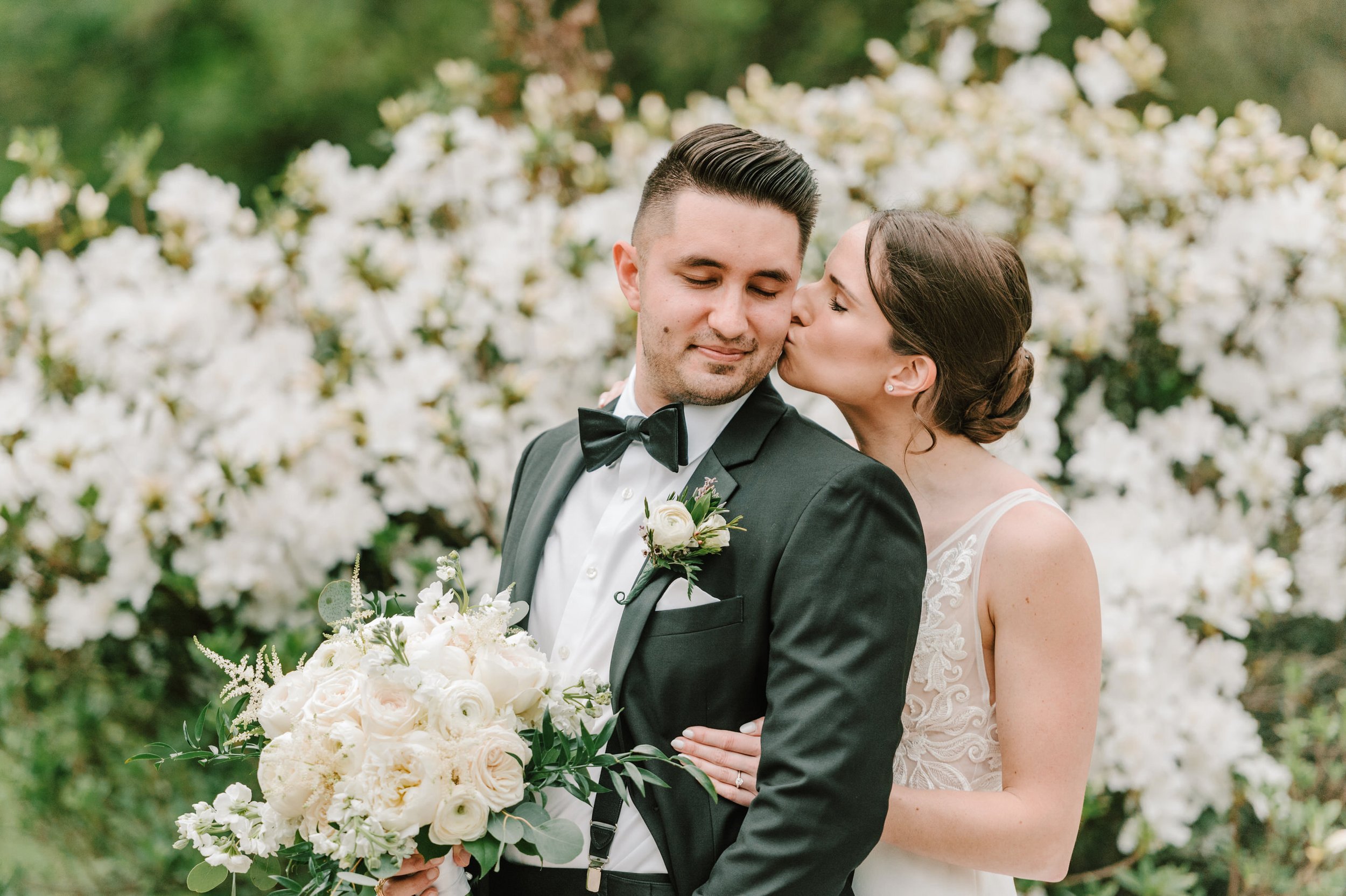 A groom and bride on their wedding day, with the bride giving the groom a kiss on the cheek. The groom is dressed in a black tuxedo with a bow tie and holding a bouquet of white flowers. The bride is in a lace wedding dress, embracing the groom from 