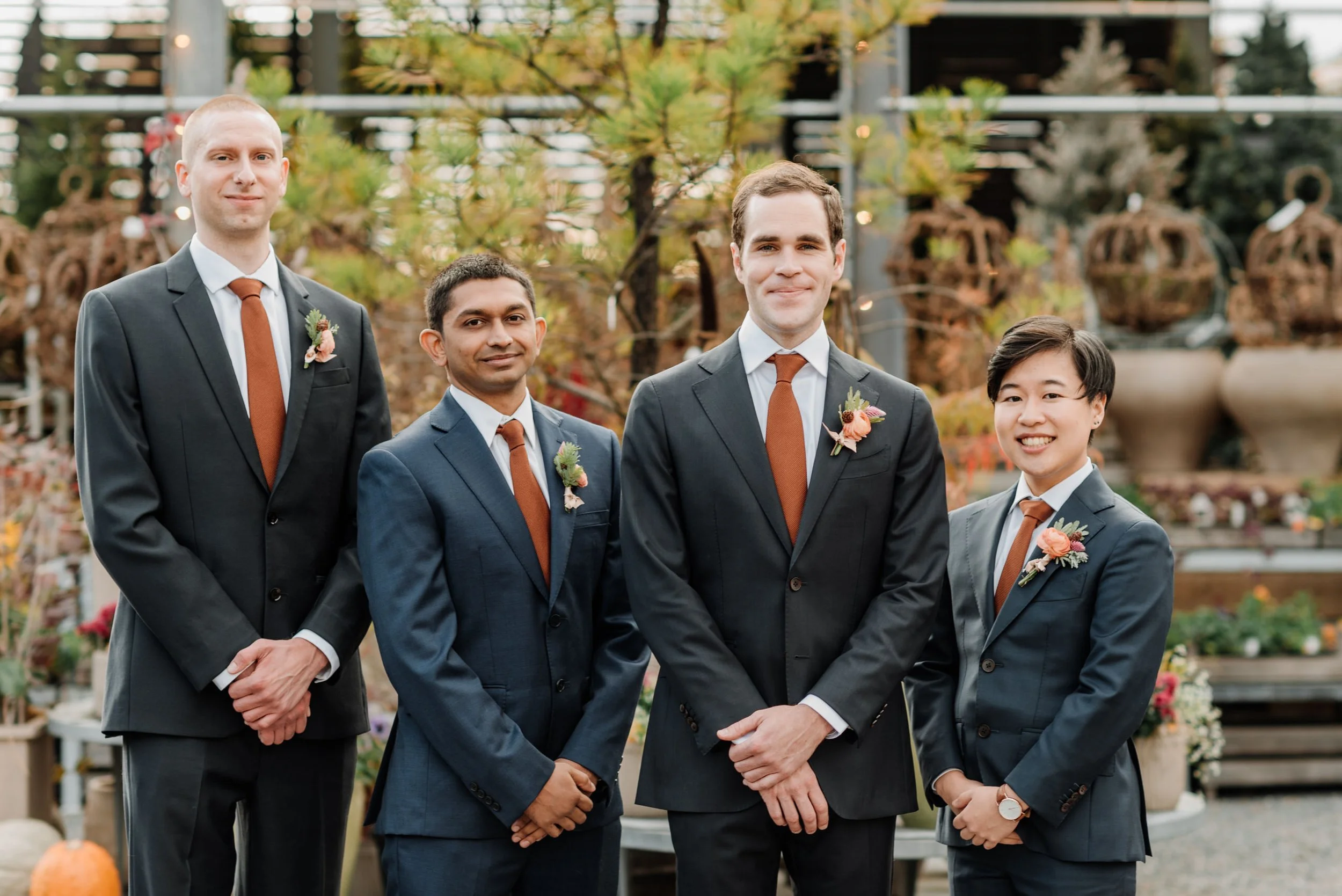 Group of five men in suits with boutonnières, standing outdoors in autumn setting with plants and trees in the background.
