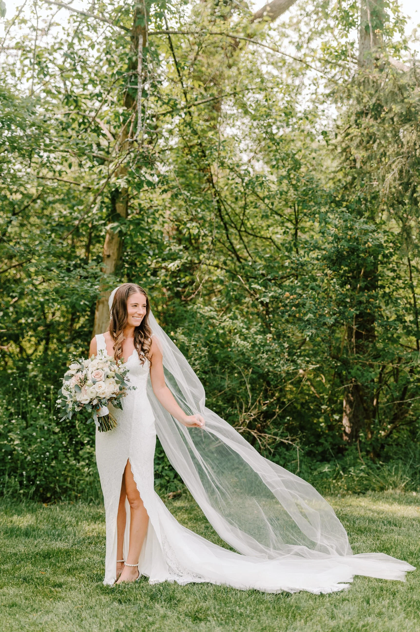 A bride in a white wedding dress holding a bouquet, standing outdoors on grass with trees in the background, smiling, and holding her long veil.