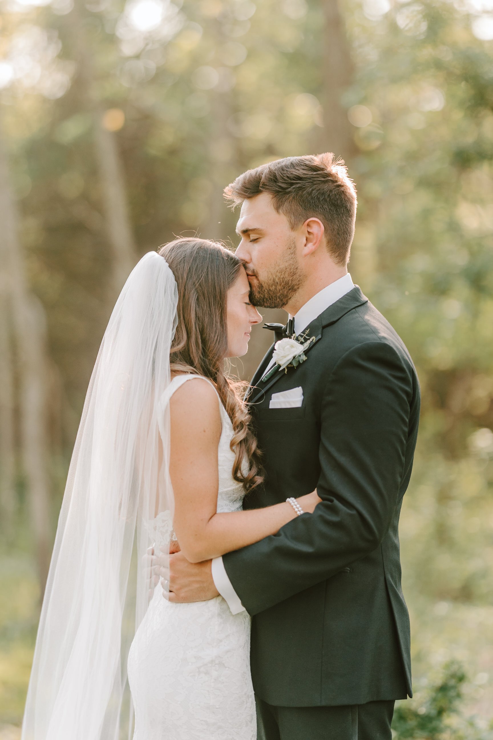 Bride and groom sharing a tender moment outdoors, with the groom kissing the bride's forehead, during their wedding.