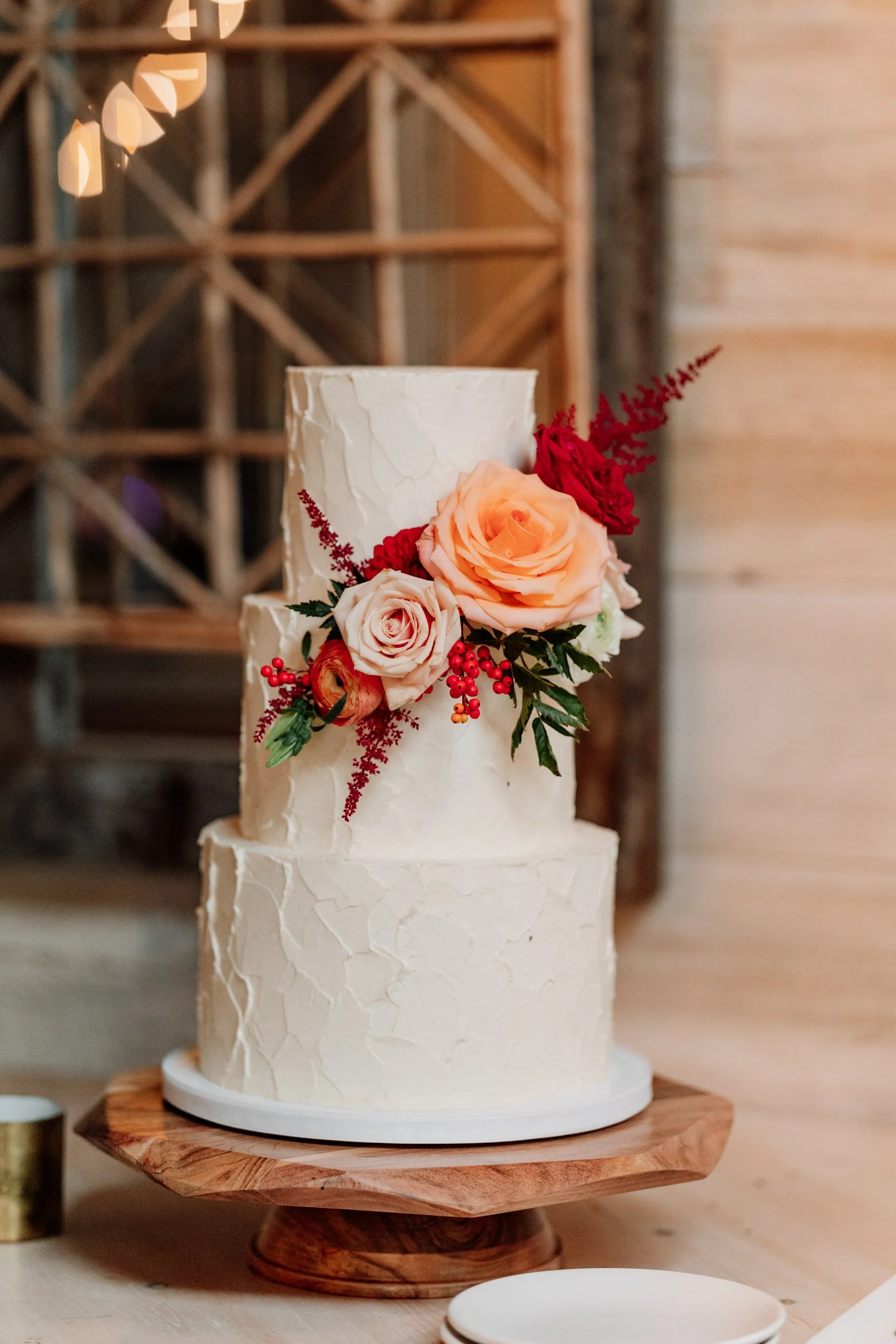 Three-tier white wedding cake with textured icing and a floral arrangement of peach, white, and red roses, along with red berries and greenery, on a wooden cake stand.