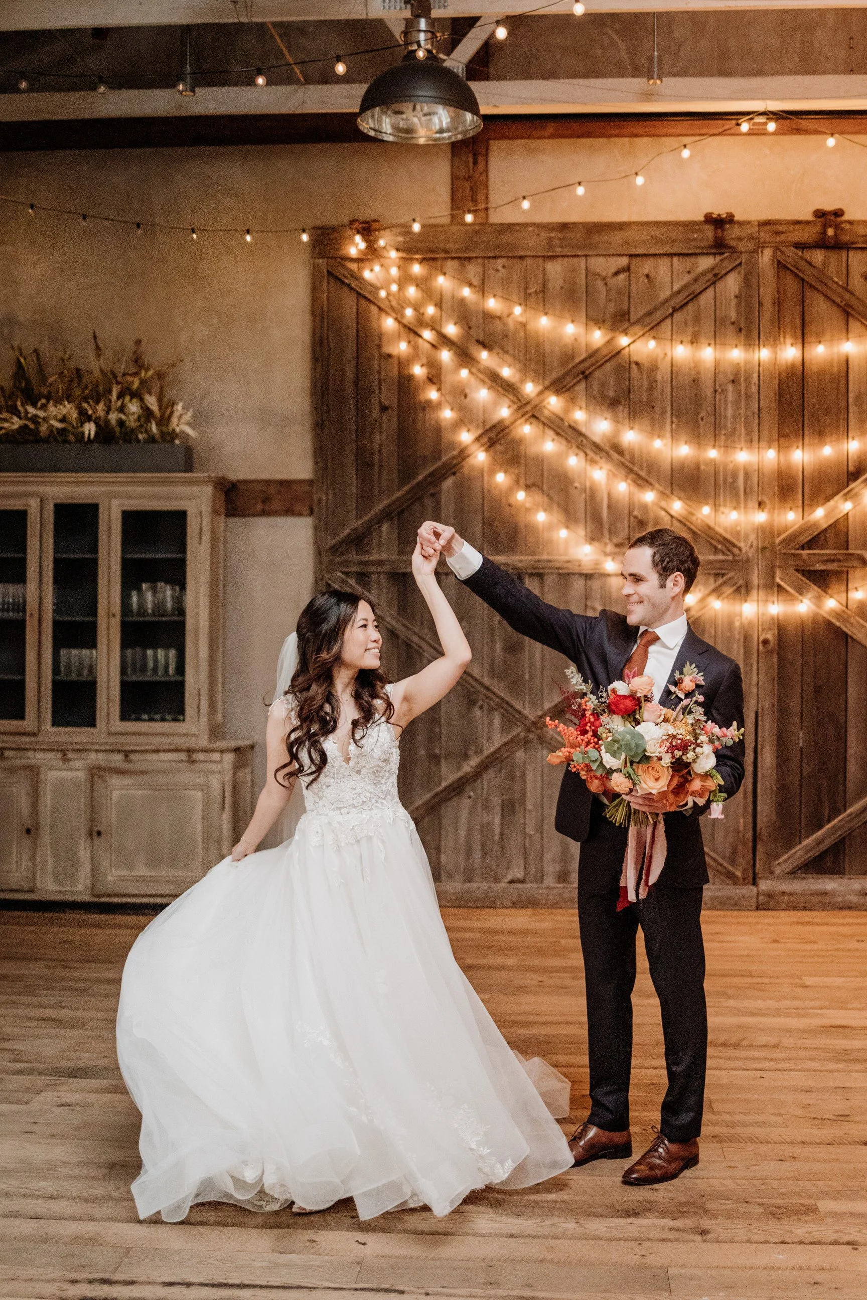 A bride and groom dancing at their wedding reception with string lights and a wooden barn door in the background.