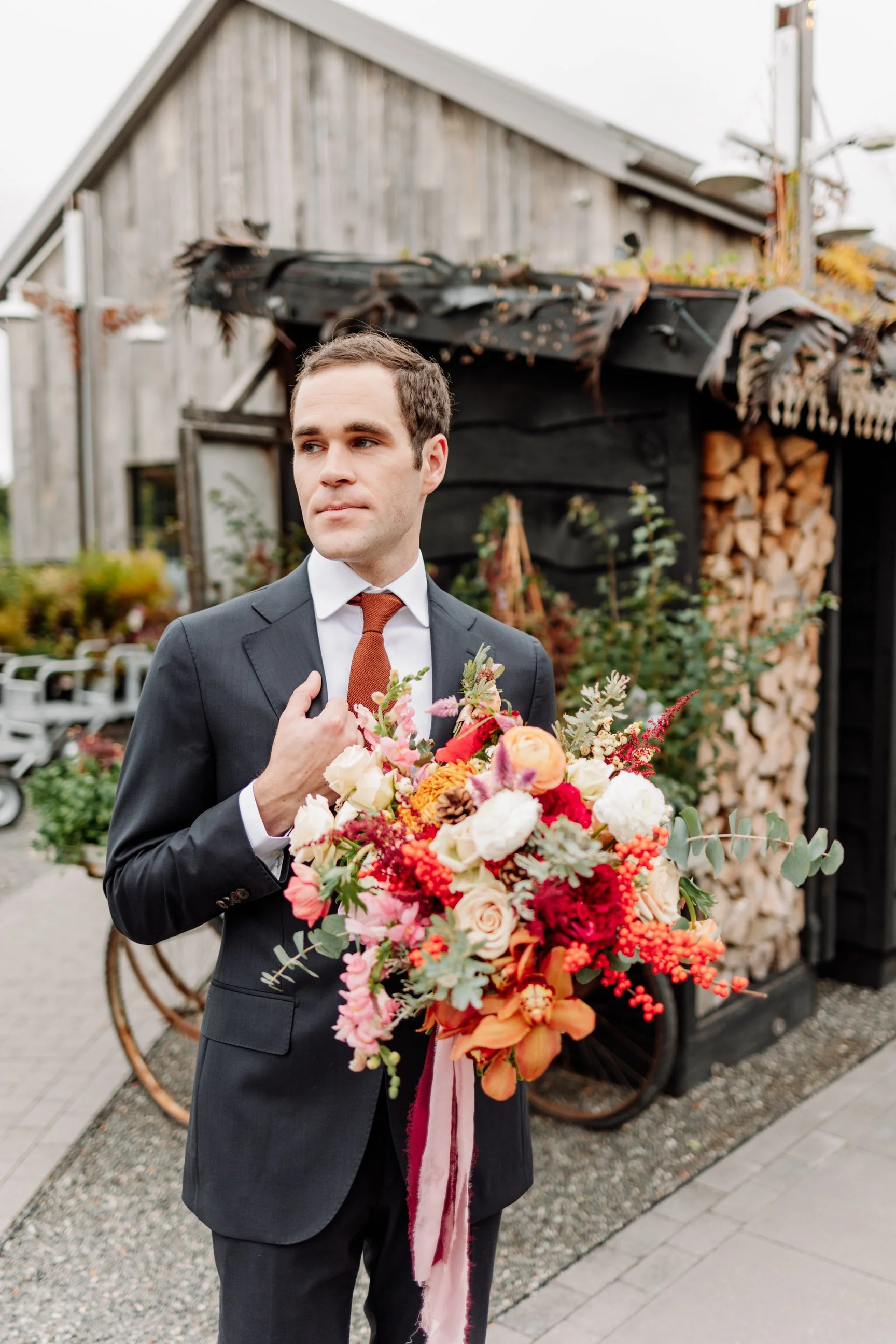 Man in a suit holding a large colorful bouquet of flowers outdoors near a rustic wood structure.