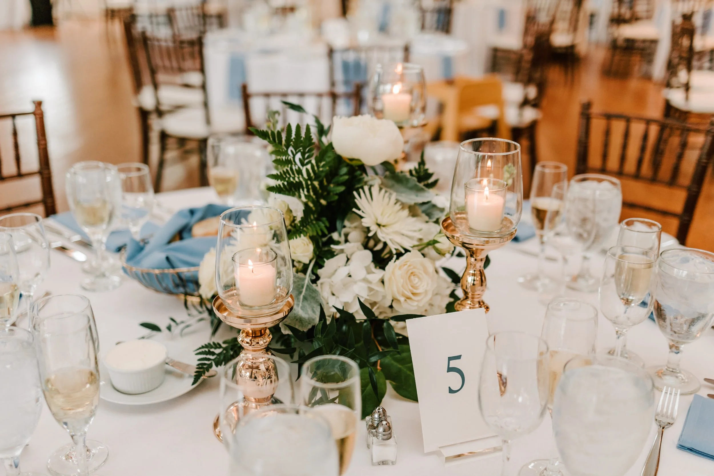 Elegant banquet table decorated with white and green floral centerpiece and candles in glass holders, with a blue napkin and table number five.