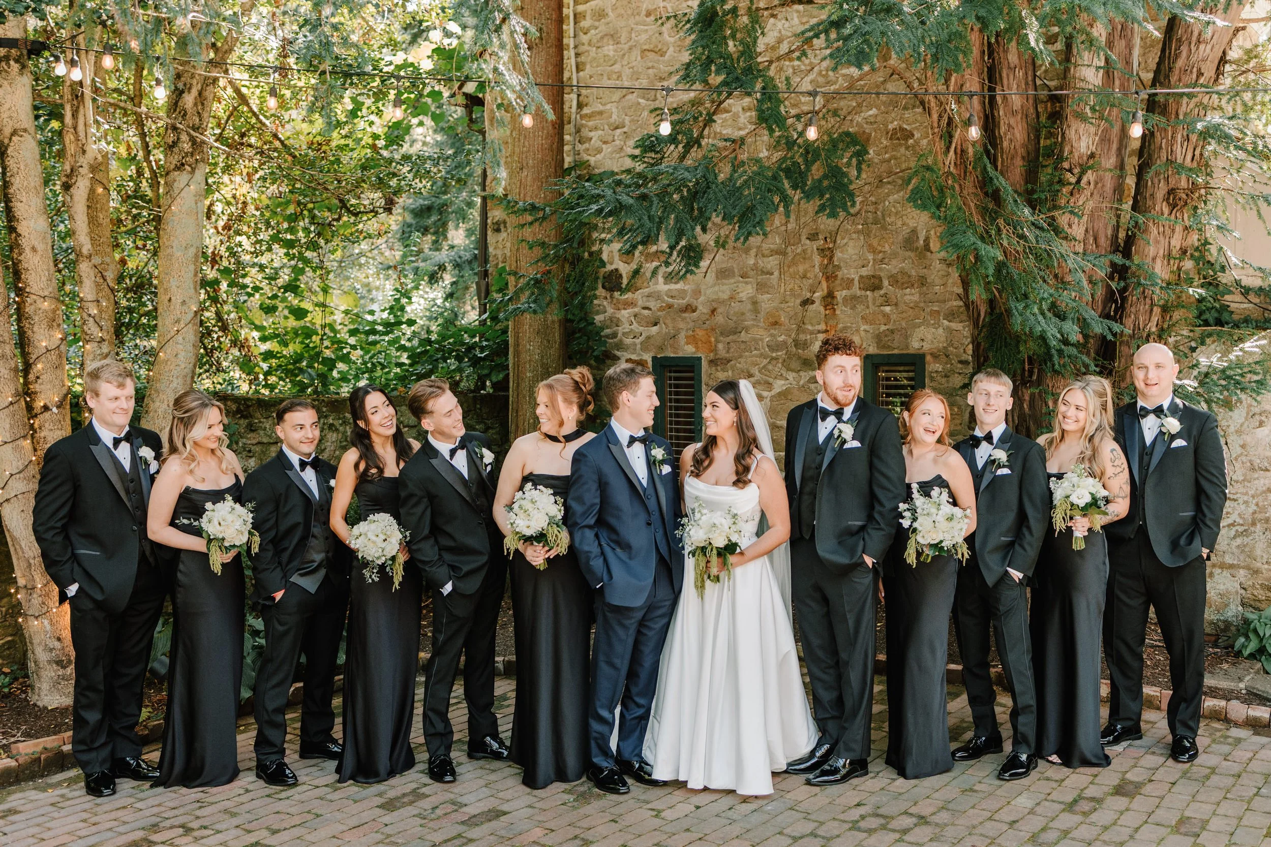 A wedding party of 14 people, including bride and groom, posing outdoors with brick and stone wall, trees, and string lights in the background.