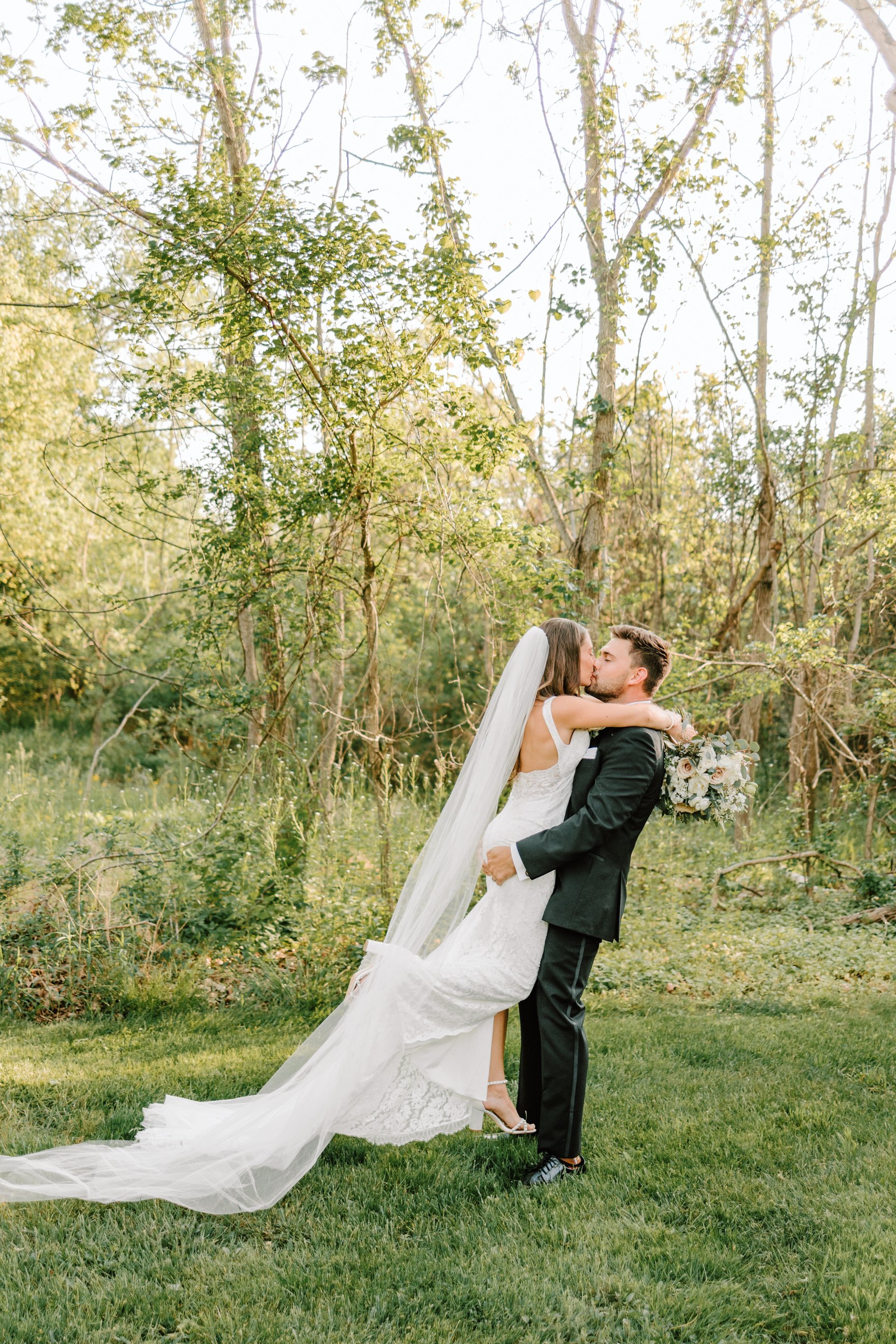 A bride and groom share a kiss outdoors in a wooded area, with the bride in a white wedding gown and veil, and the groom in a black suit, as she lifts him slightly off the ground.