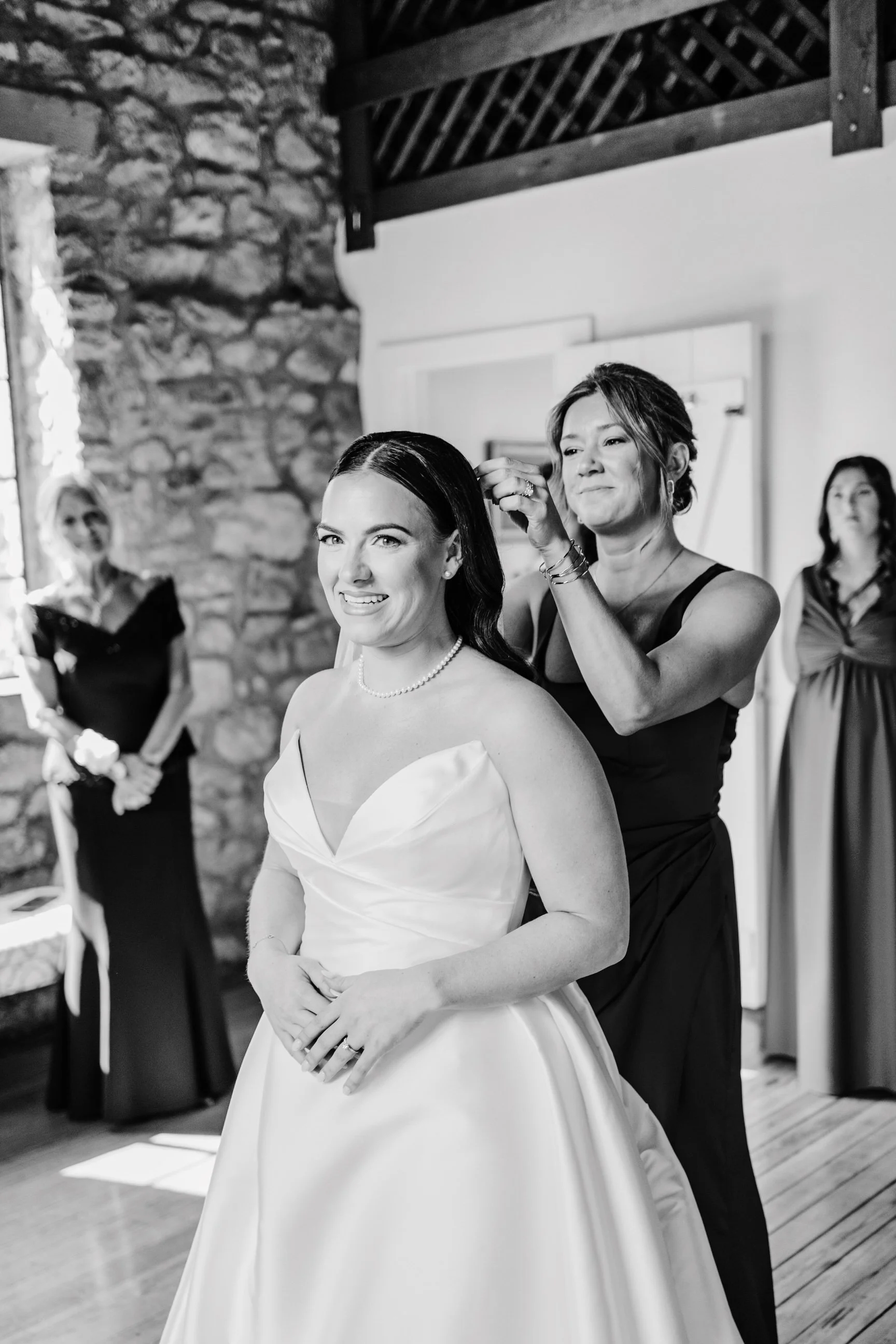 A bride with dark hair in a white strapless wedding gown sits while a woman assists her with her hair during a wedding preparation. Two other women observe in the background, one holding a bouquet, inside a rustic room with a stone wall and wooden ce