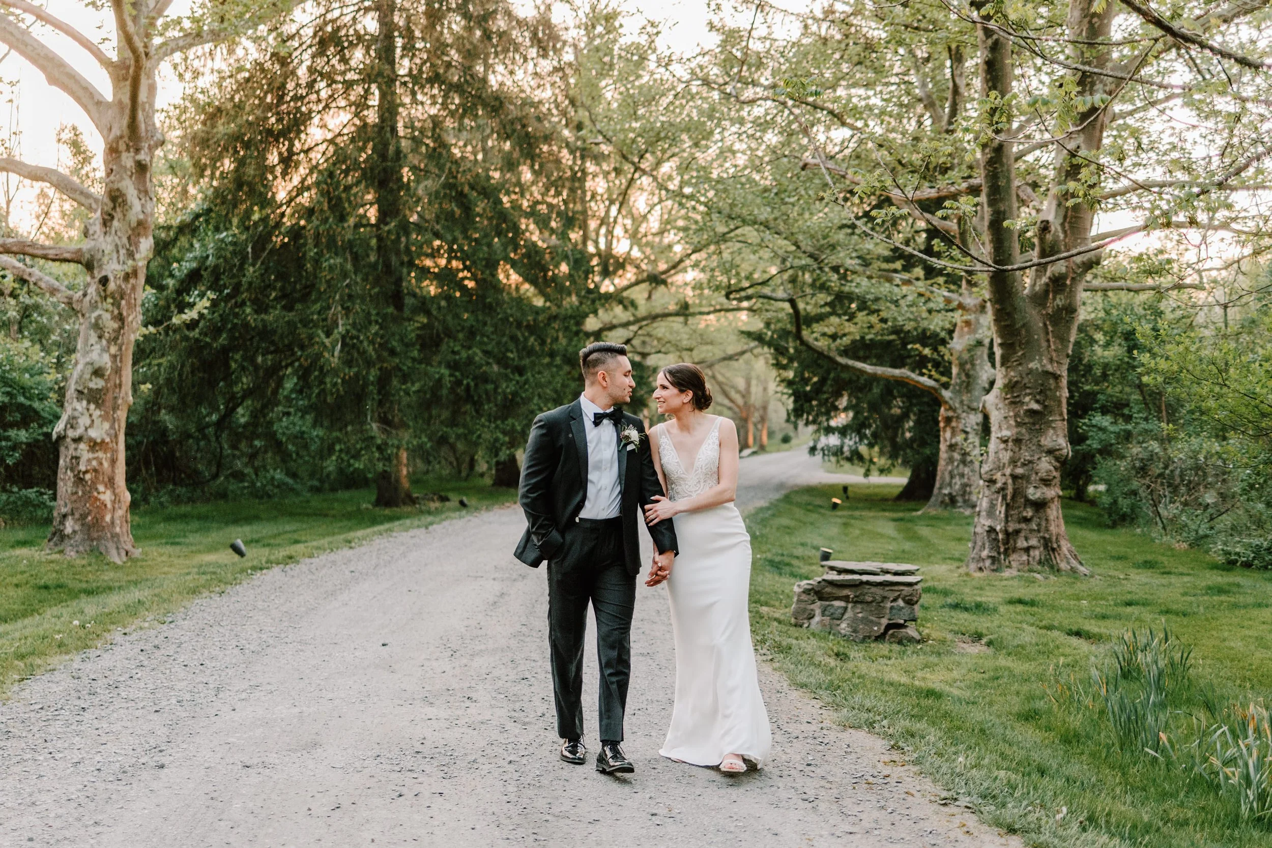 A newlywed couple walking hand in hand along a gravel park path surrounded by trees at sunset.