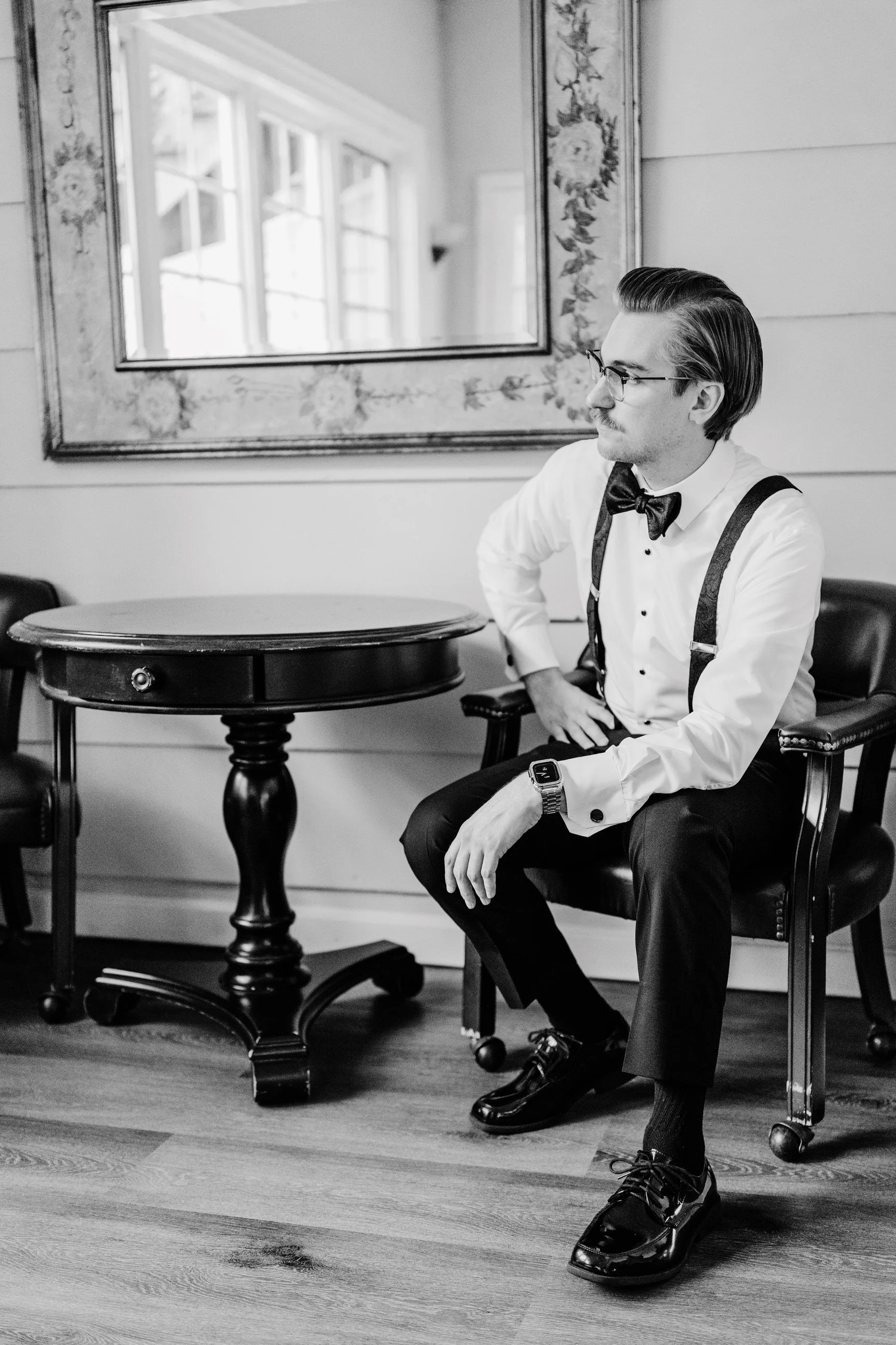 A man dressed in formal attire, including a tuxedo shirt, bow tie, suspenders, and shiny dress shoes, sits on a black upholstered chair in a room with a wooden floor, a round wooden table, and a large mirror on the wall.