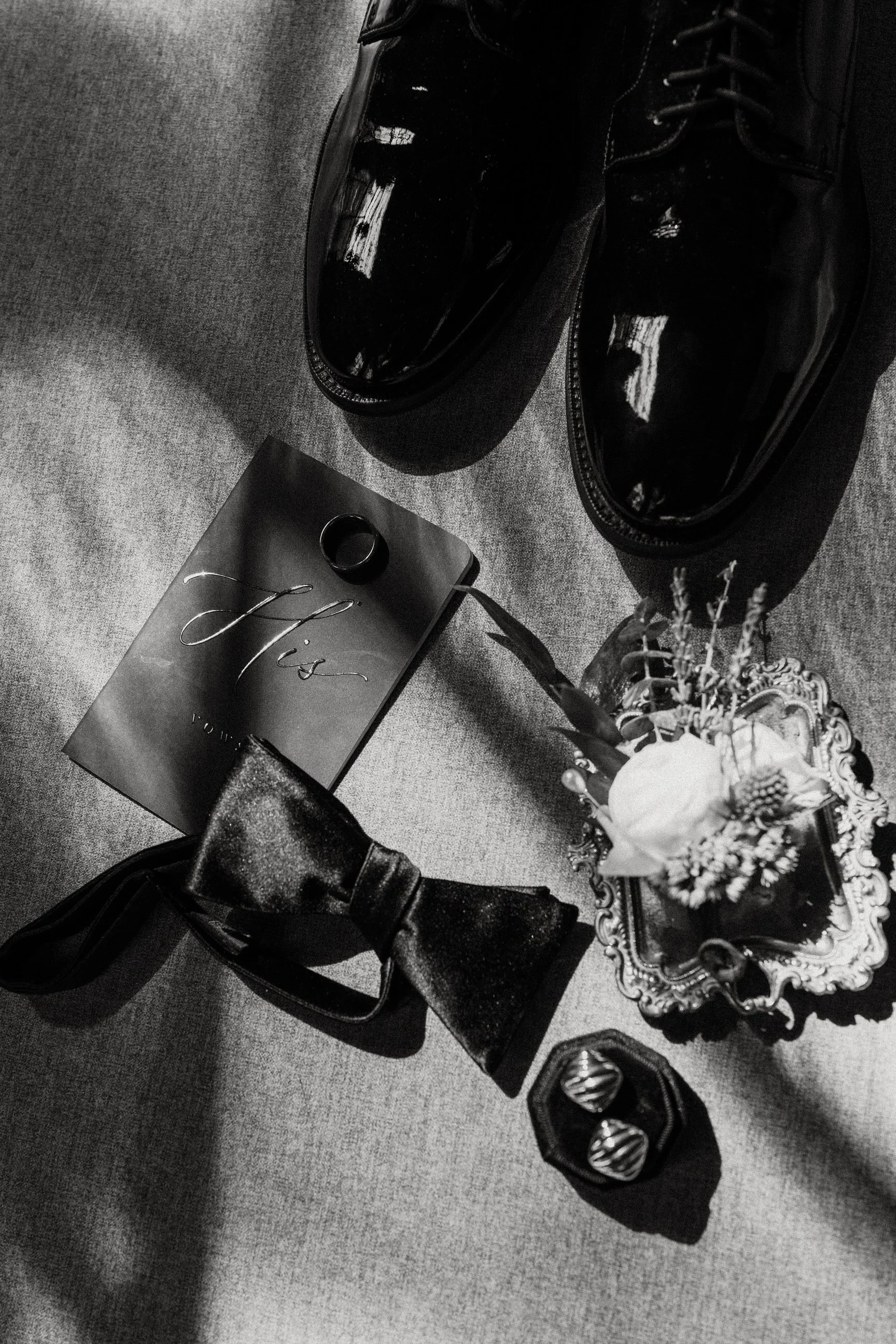 Black patent leather shoes, a flower in a decorative frame, a bow tie, a ring, and a pair of cufflinks on a fabric surface.