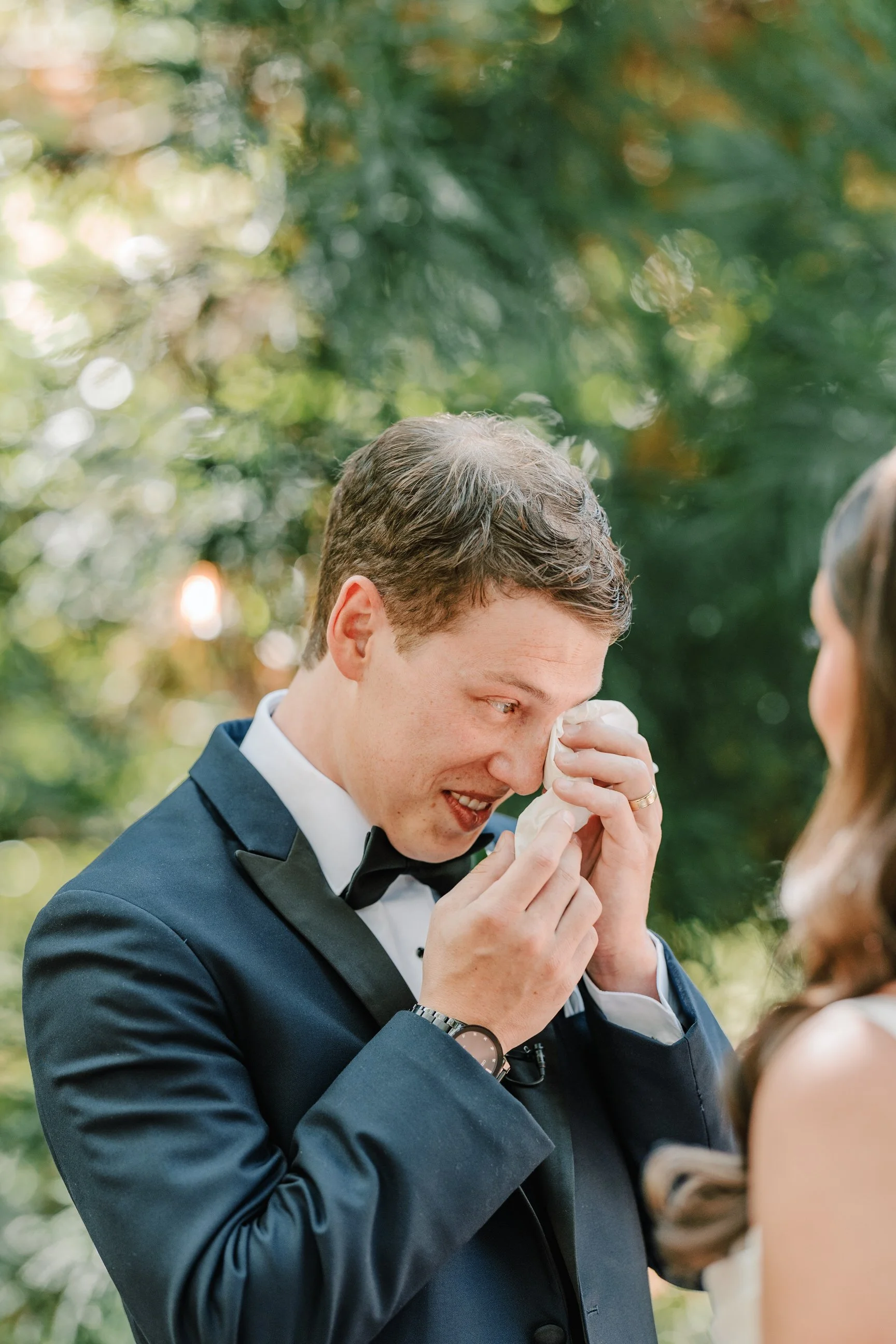 A man in a tuxedo wiping away tears at an outdoor wedding ceremony.