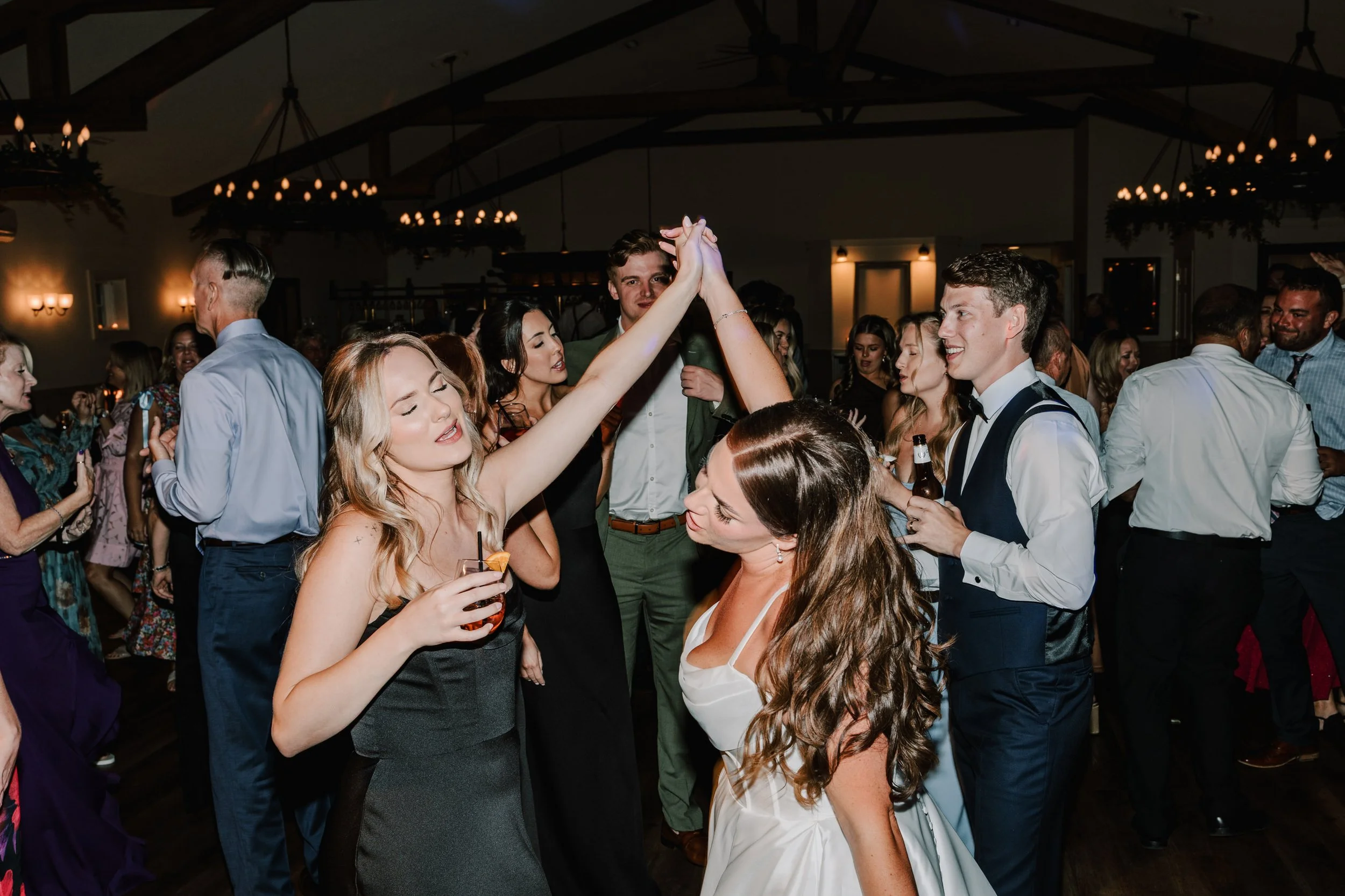 People dancing and socializing at a party or wedding reception, with a woman in a black dress and a woman in a white dress holding hands and dancing.