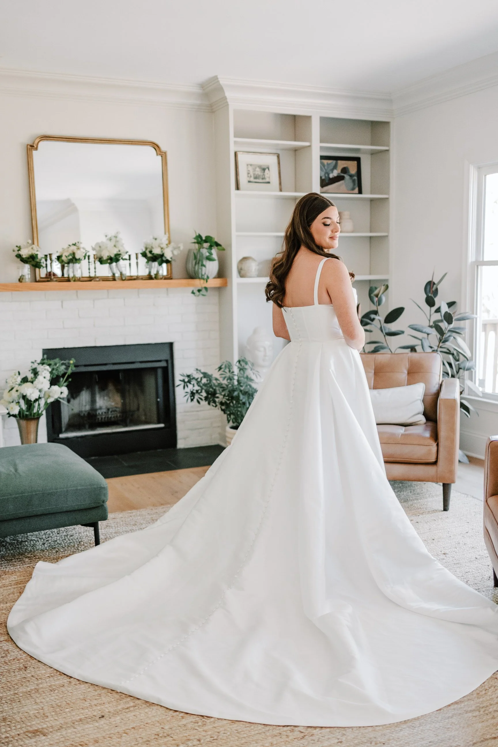 A woman in a white wedding gown standing in a bright, cozy living room with a fireplace, plants, and built-in shelves.