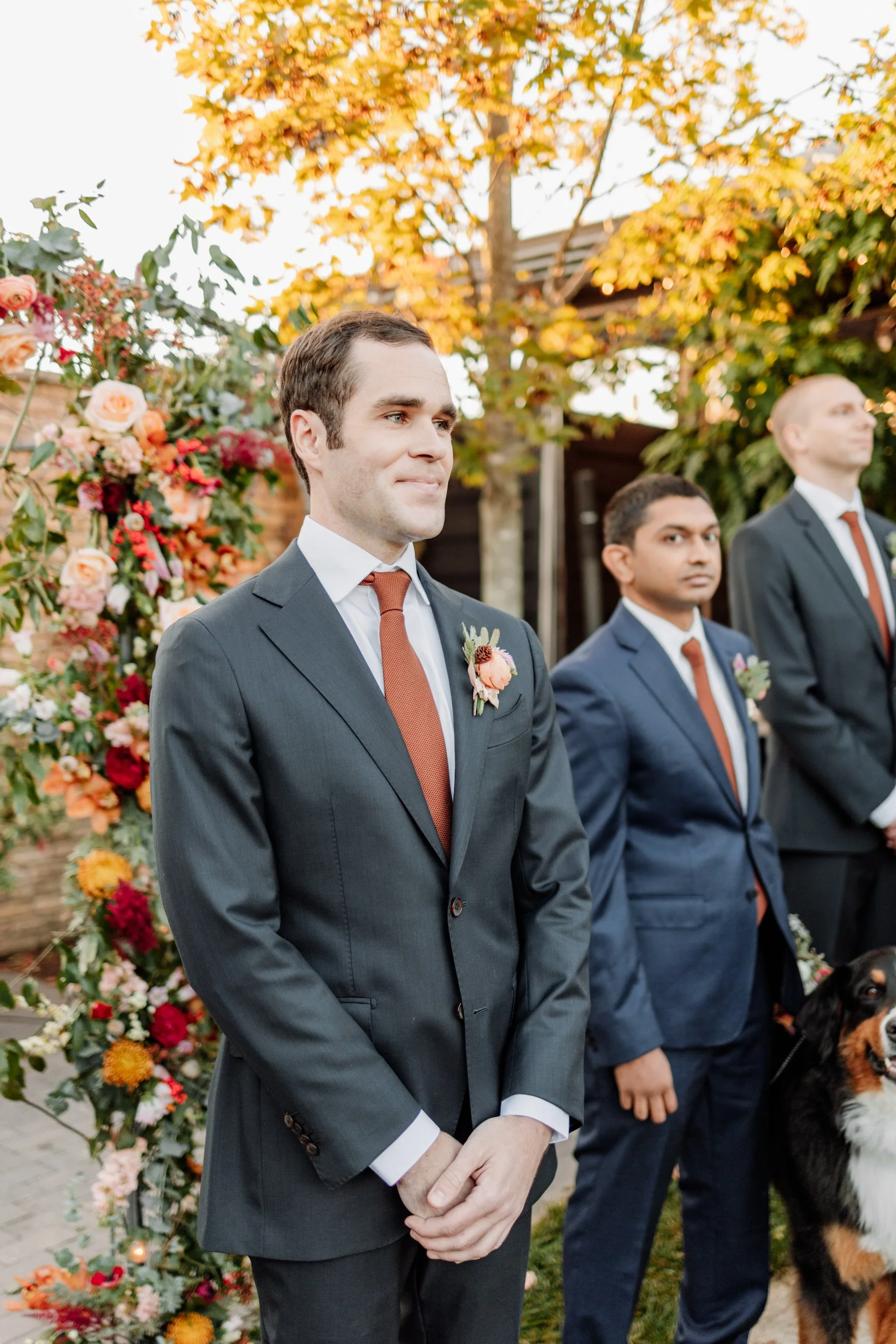 A groom in a dark suit with a brown tie and a boutonniere, standing outdoors during a wedding ceremony, with fall foliage in the background and other groomsmen along with a dog nearby.