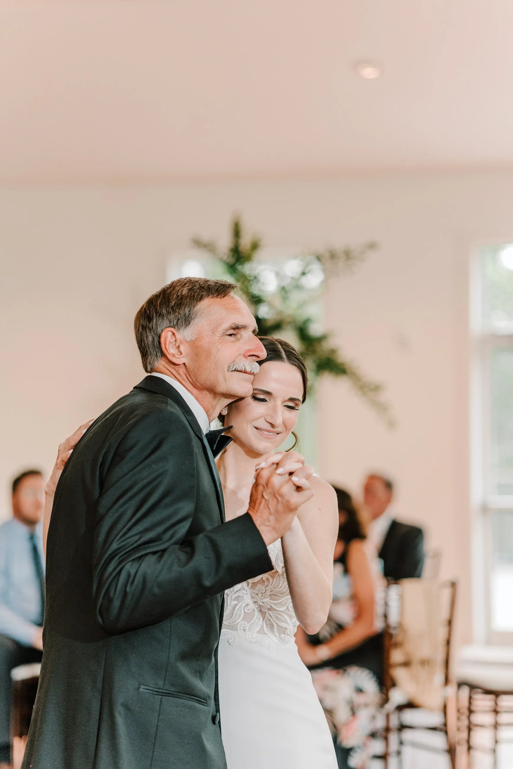 A father and daughter share a dance at her wedding, smiling with eyes closed, in a bright reception hall decorated with greenery and a star-shaped decoration in the background.