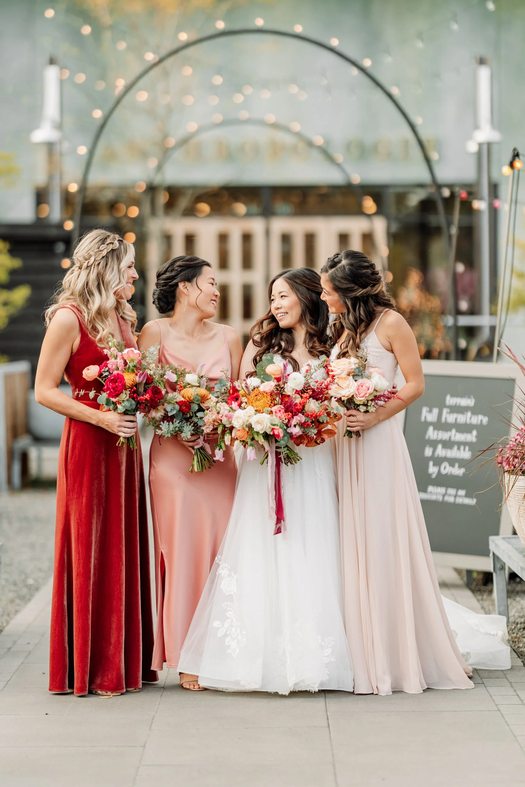 Four women in formal dresses holding bouquets of flowers, standing together outdoors during a festive event, with fairy lights overhead and a decorative backdrop.