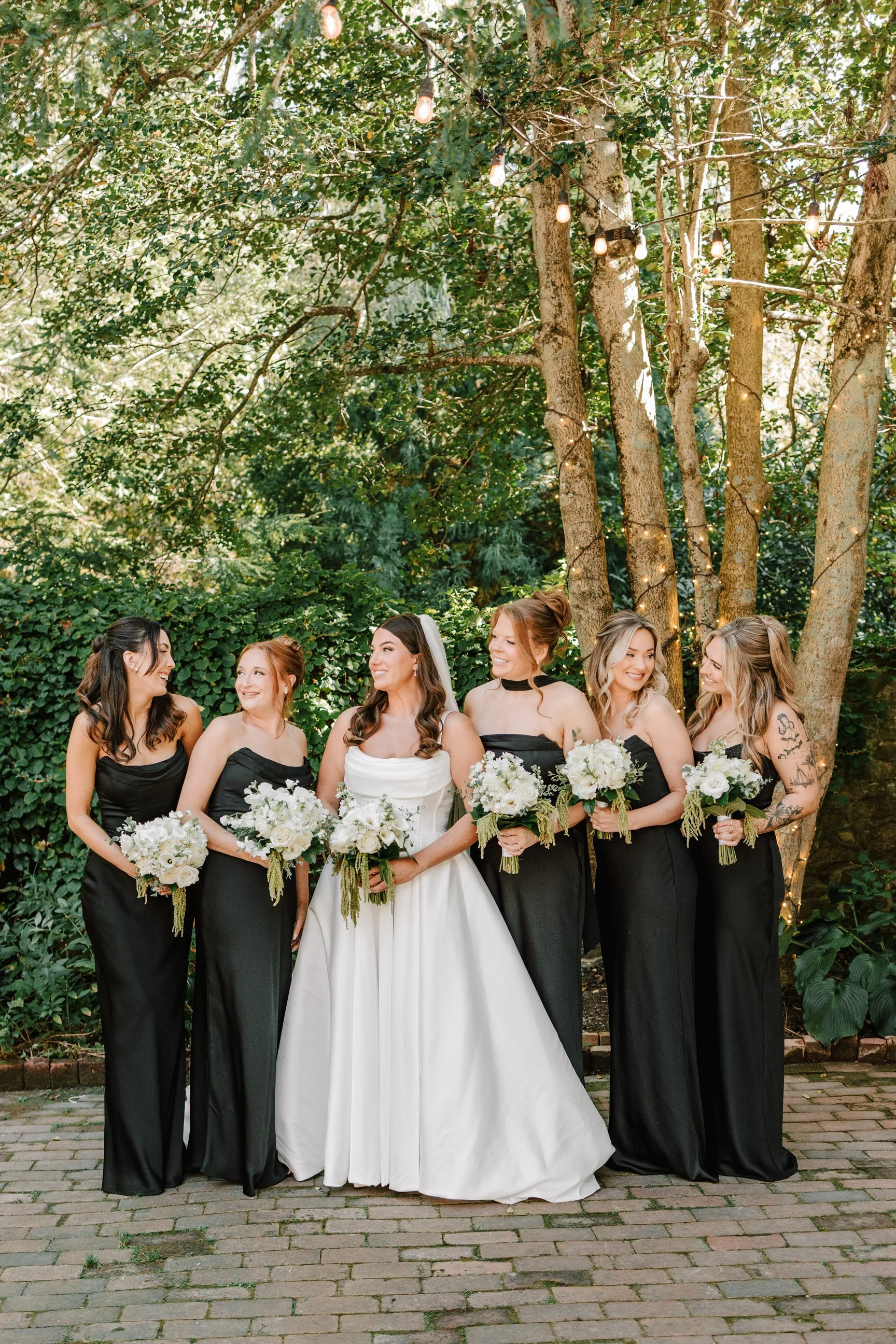 A group of six women, including a bride in a white wedding gown with a veil, and five bridesmaids in black dresses, standing outdoors on a brick pathway under trees with string lights.
