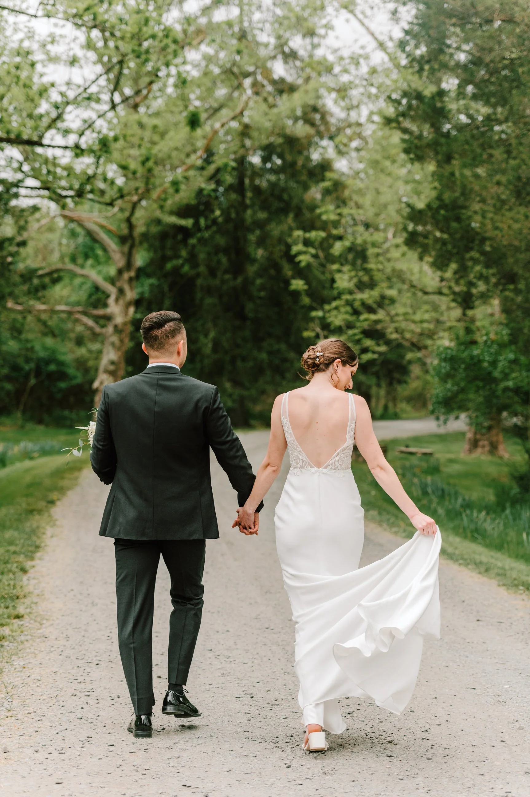 A bride and groom walking hand in hand on a tree-lined dirt path, with the bride holding up her wedding dress.