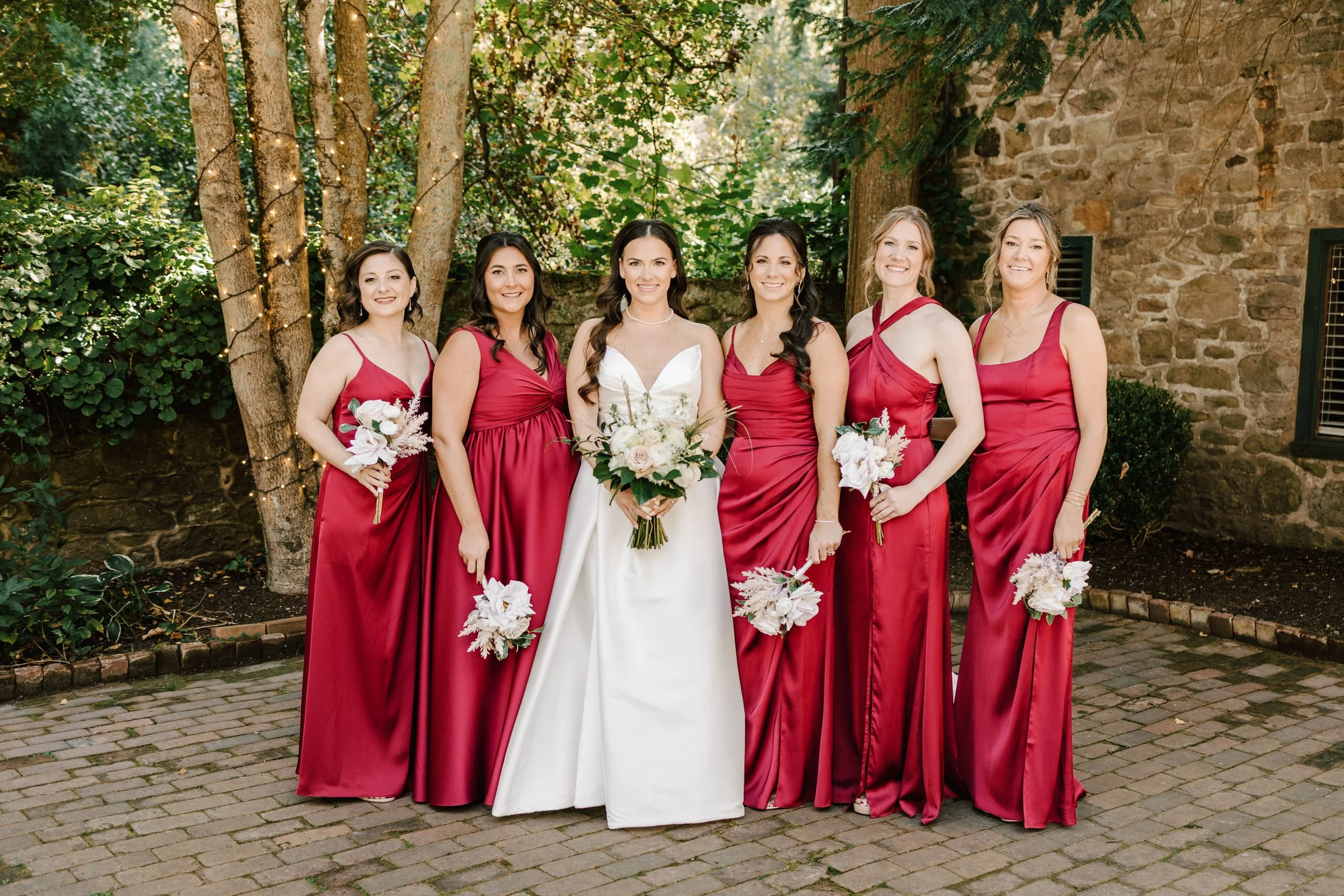 A bride in a white wedding dress standing with six bridesmaids in matching red dresses, all holding bouquets, outdoors in front of trees and a stone wall.