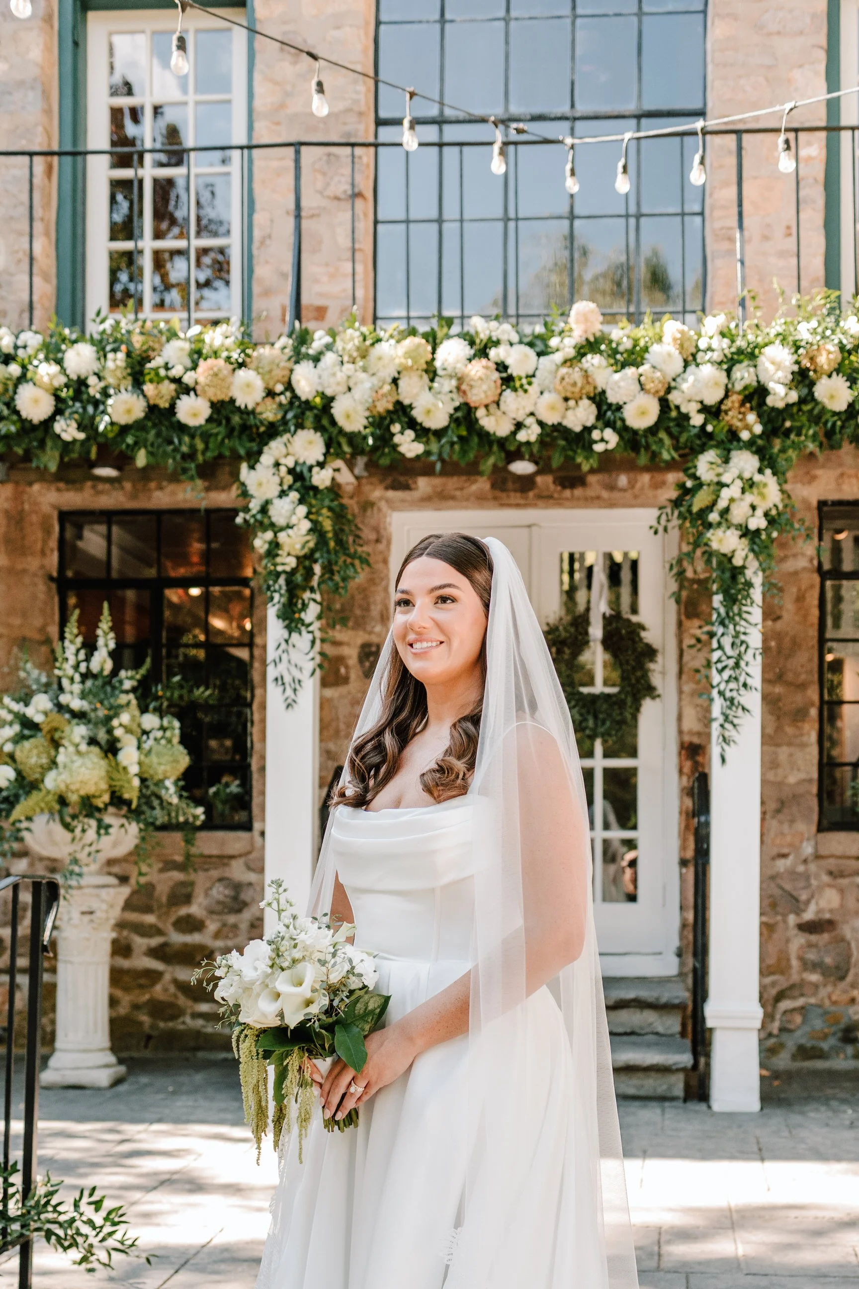 A bride in a white wedding dress holding a bouquet walking outside in front of a floral arch with white and blush flowers, under string lights, in front of a stone building with large windows.