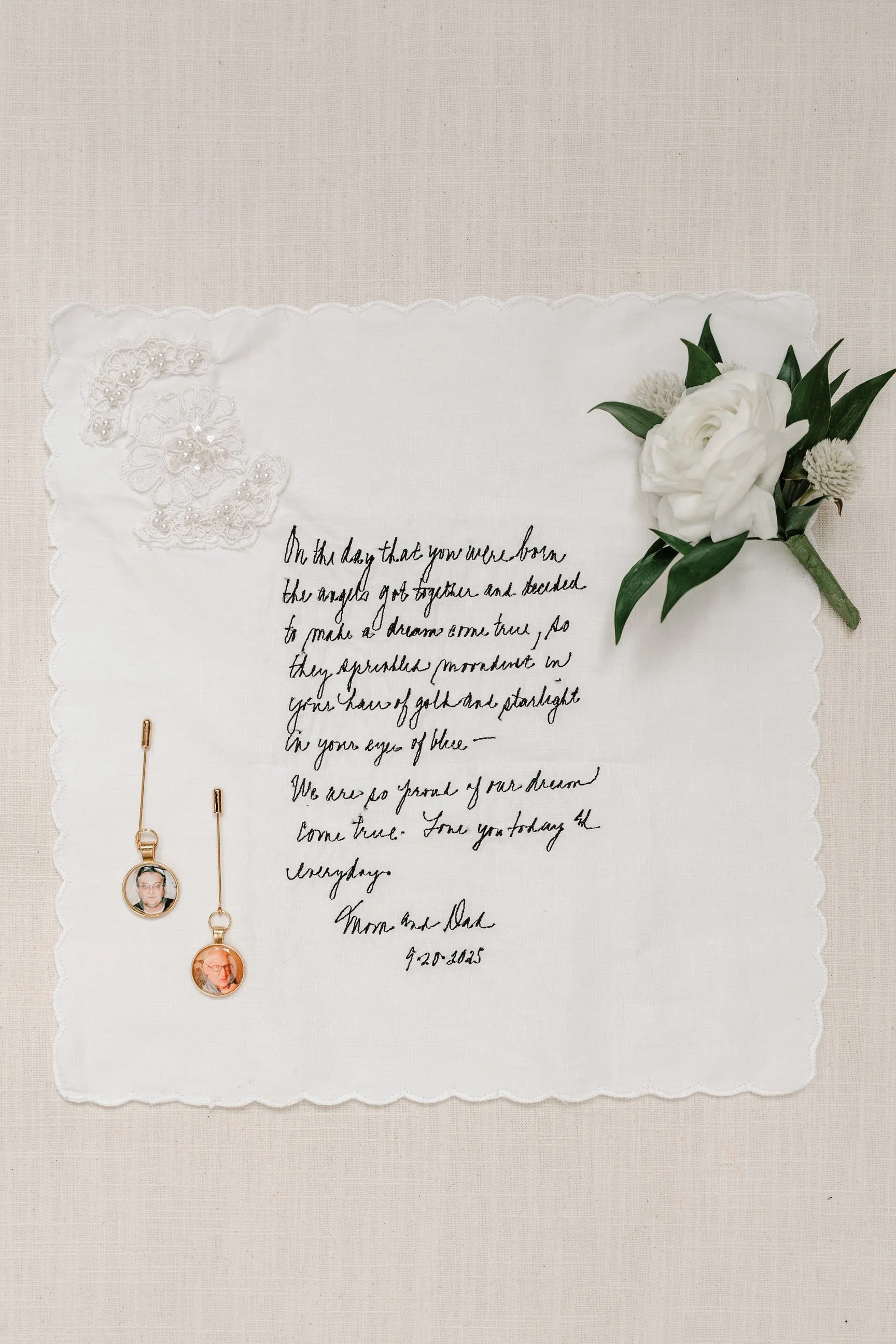 A wedding keepsake with a handwritten poem, a white flower boutonniere, and a pair of gold earrings with photo charms, all on a white scalloped cloth. The poem is signed by the couple with a date.