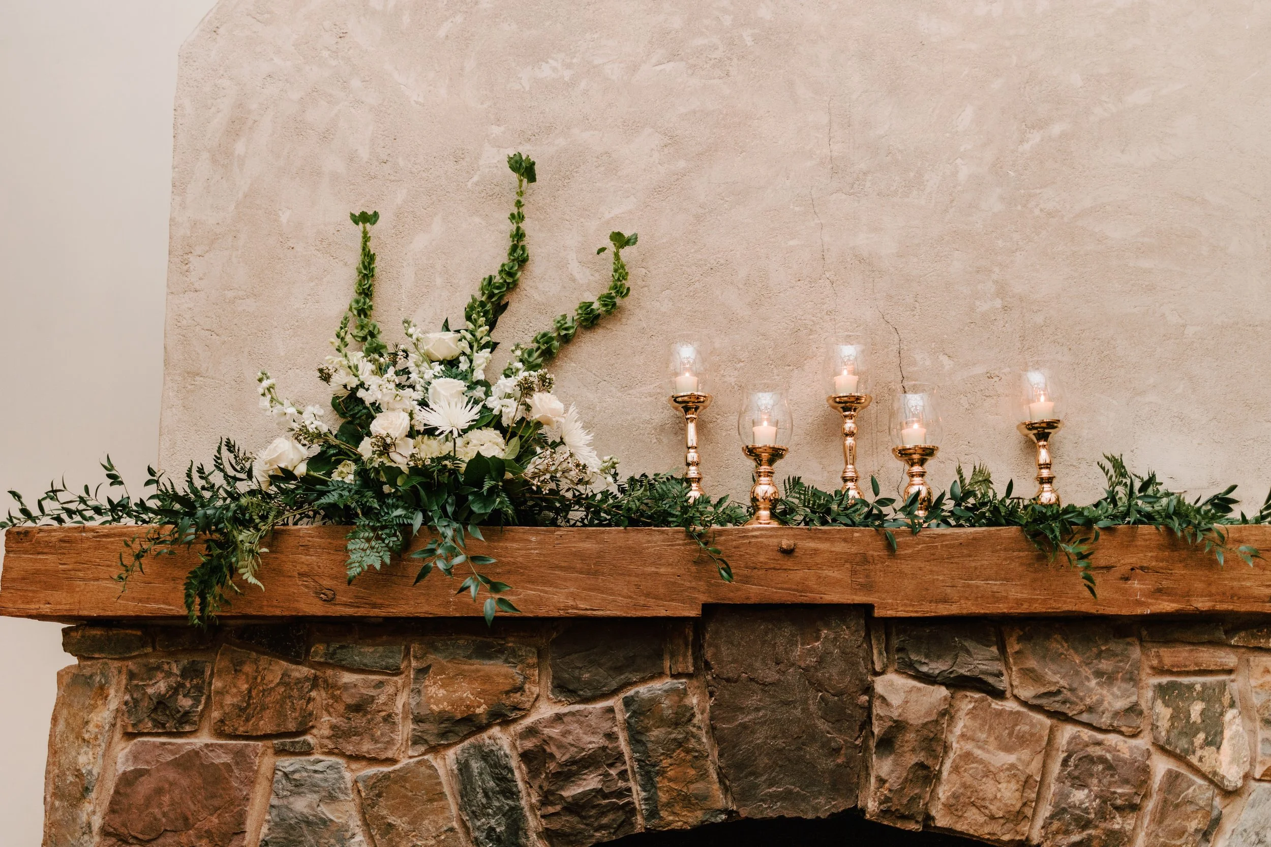 Decorative arrangement on a wooden mantel above a stone fireplace, featuring white flowers, greenery, and five lit candles in bronze holders