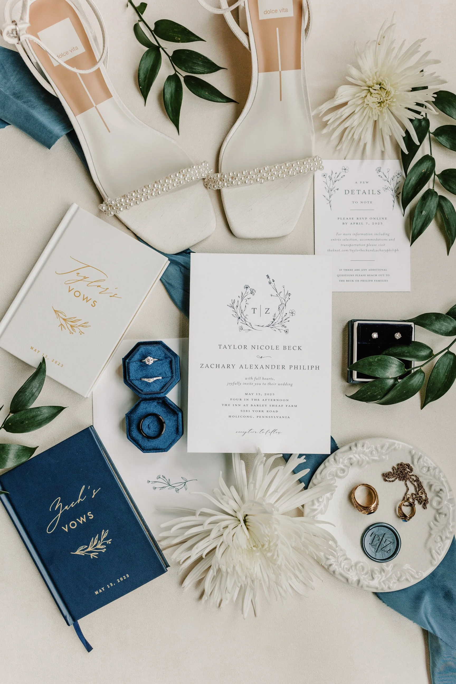 Wedding invitation, engagement ring, wedding rings, jewelry, white flowers, green leaves, bridal shoes, vow books, and wedding details scattered on a table.