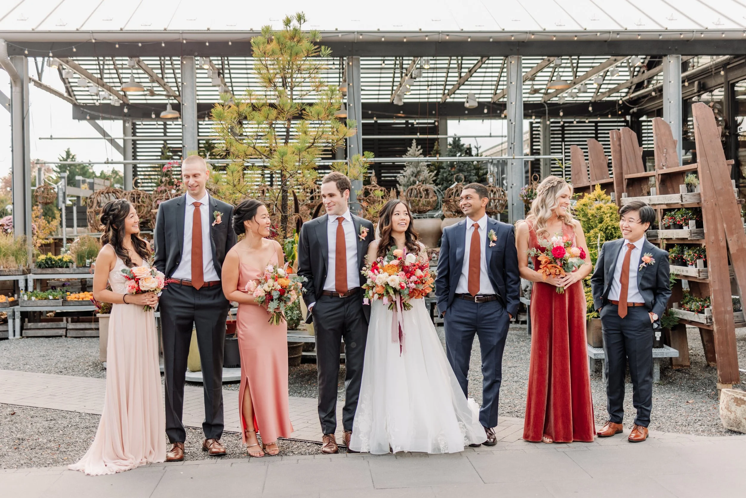 Group of eight people in formal attire, smiling and holding bouquets, in a greenhouse or garden center with plants and trees.