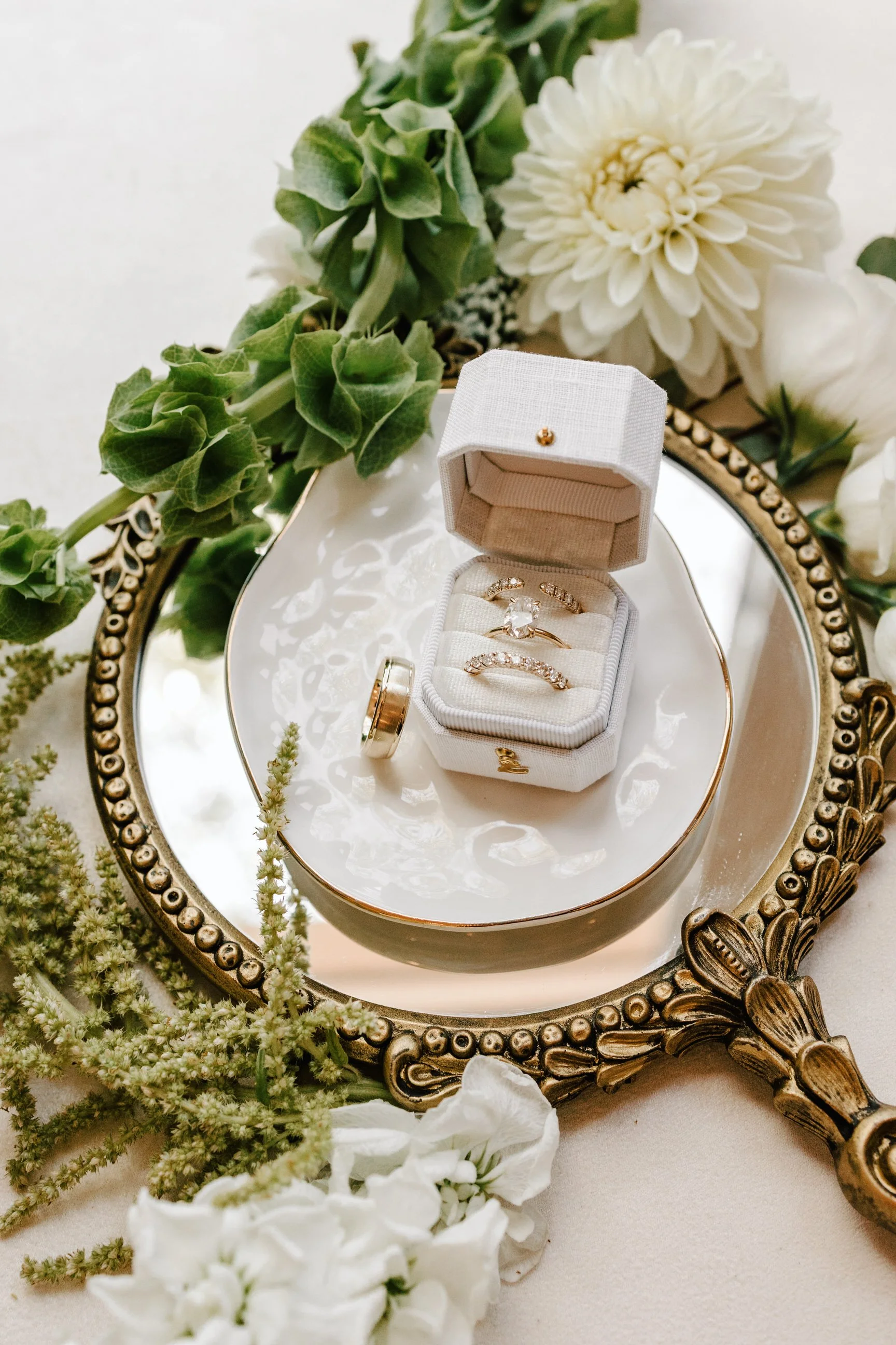 Ring box with three rings inside, surrounded by white flowers and greenery, placed on a decorative mirror
