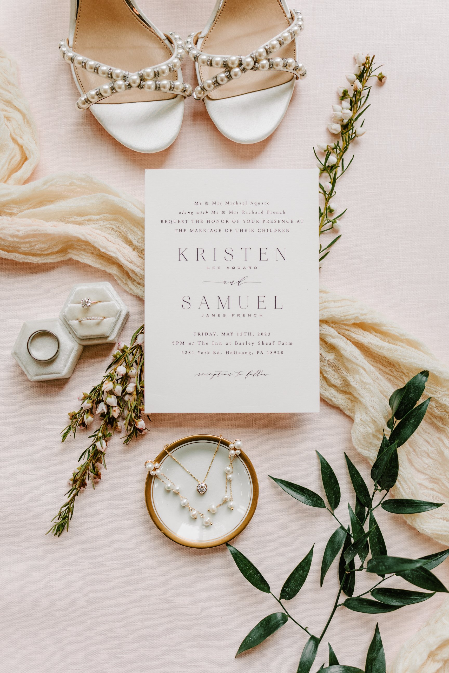 Wedding invitation surrounded by pearl sandals, jewelry, flowers, and greenery on a pink background.