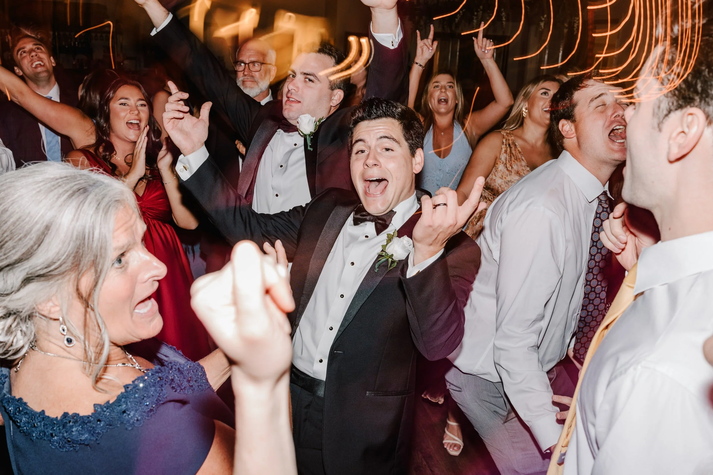 A lively wedding reception with guests dancing and celebrating. The bride and groom are in the center, dressed in formal attire, among smiling and cheering guests.
