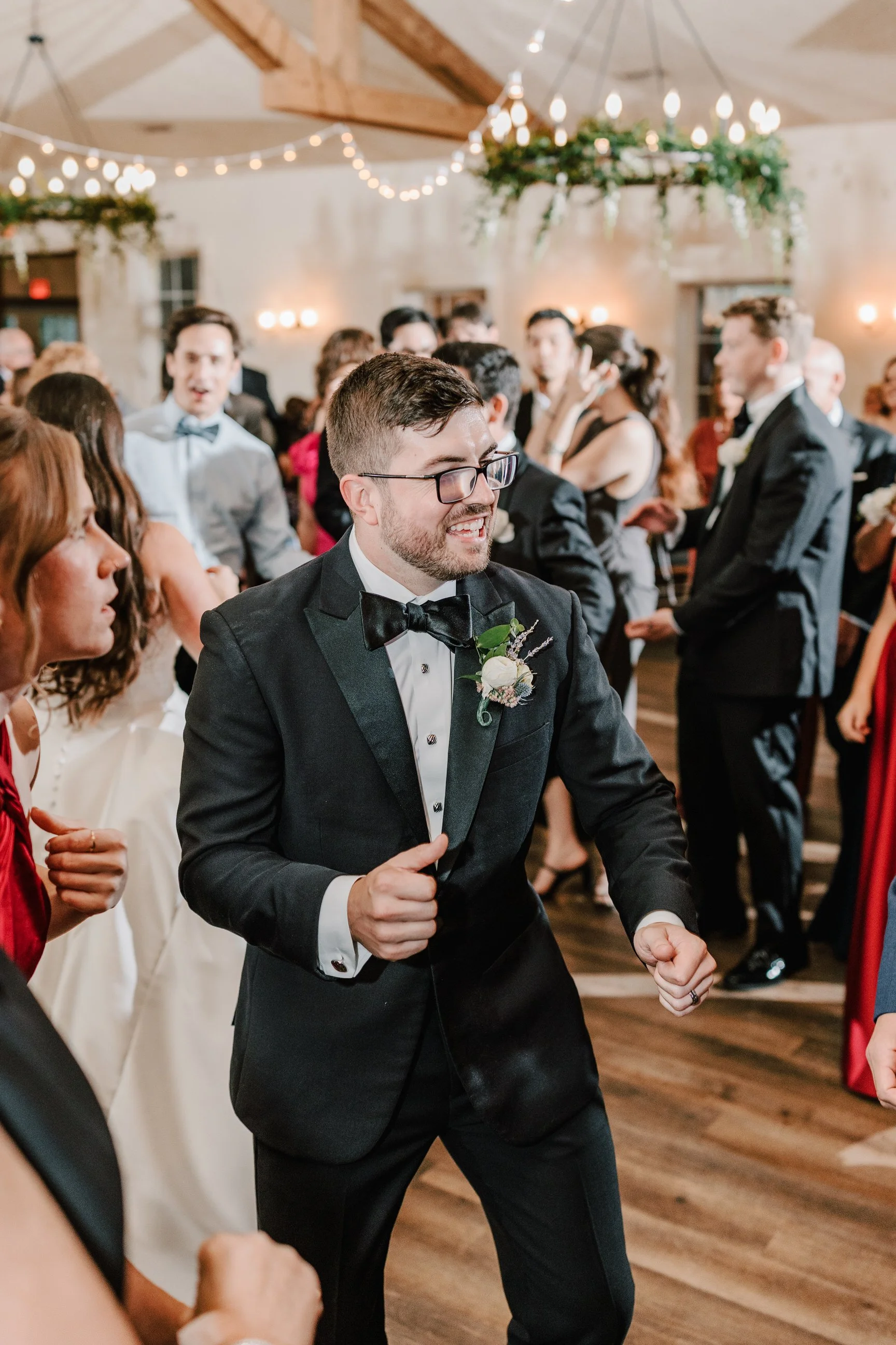 A groom dancing at a wedding reception, surrounded by guests, in a decorated venue with hanging lights and greenery.