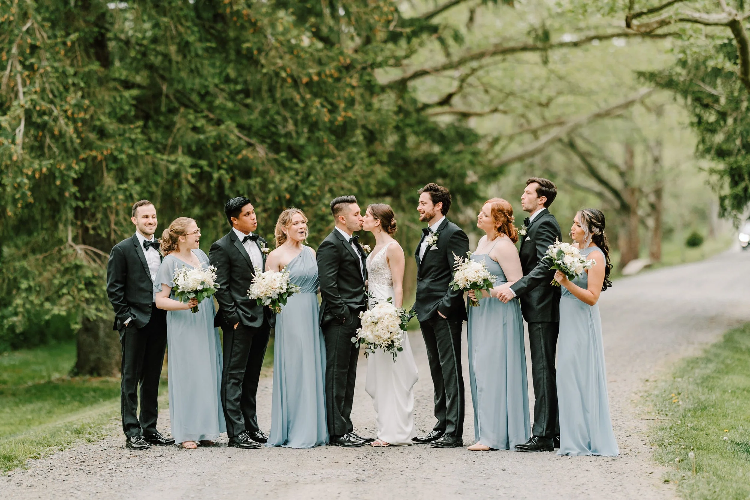 Bride and groom kissing surrounded by wedding party on a gravel path in a green park.