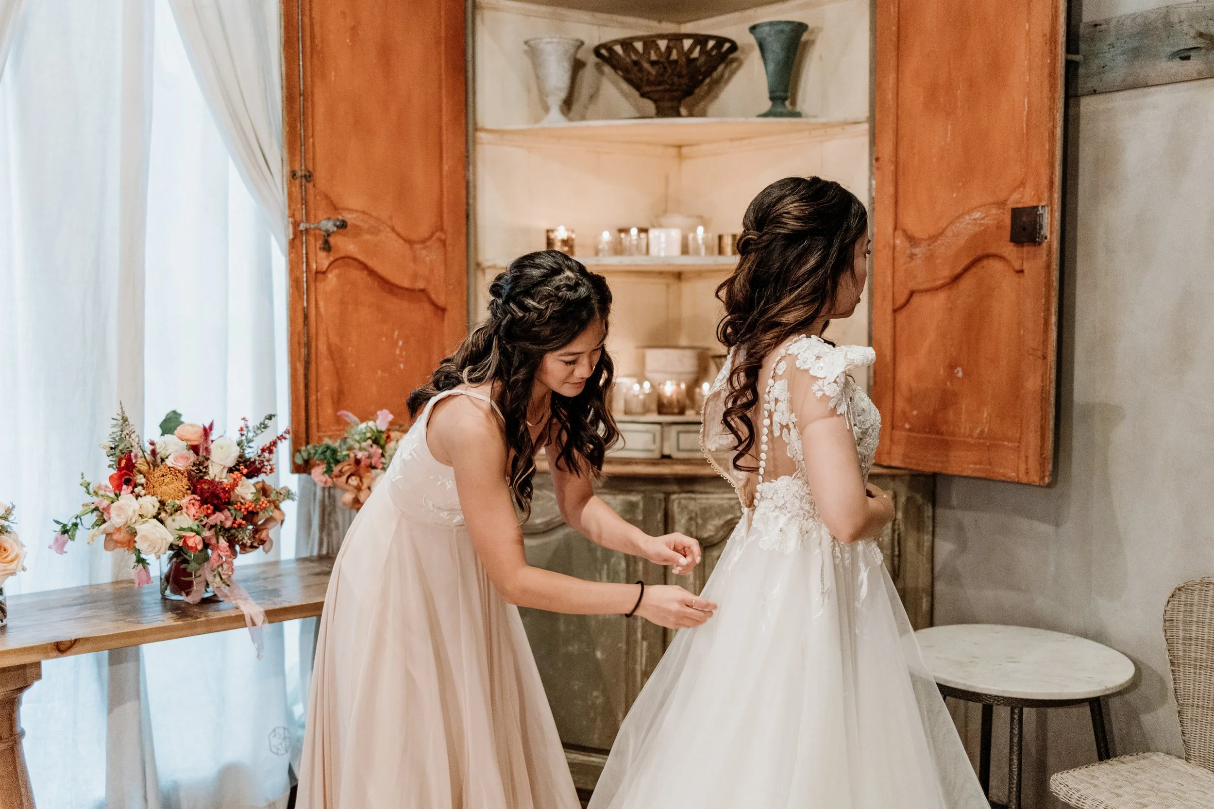 A bride in a white wedding dress with lace details is being helped into her gown by a bridesmaid in a light pink dress. The setting is a room with a wooden cabinet, floral arrangements, and candles.