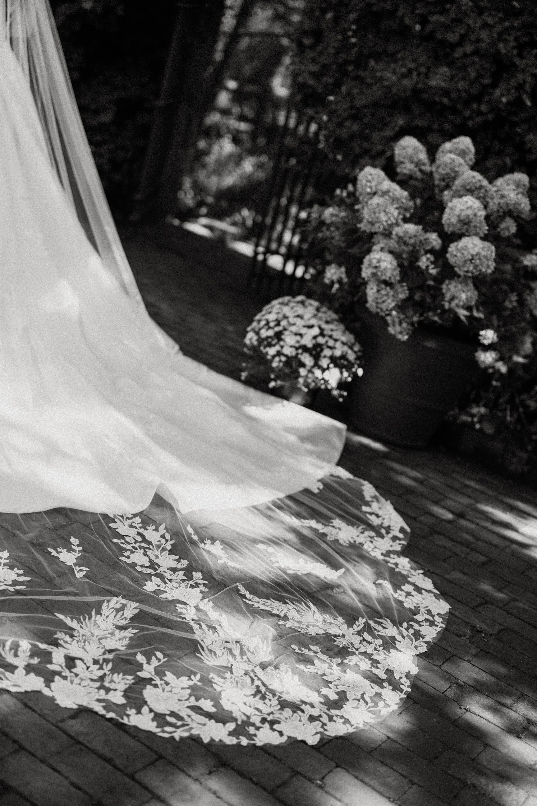 A bridal veil with intricate lace embroidery is laid on a brick floor near potted flowers and outdoor fencing.