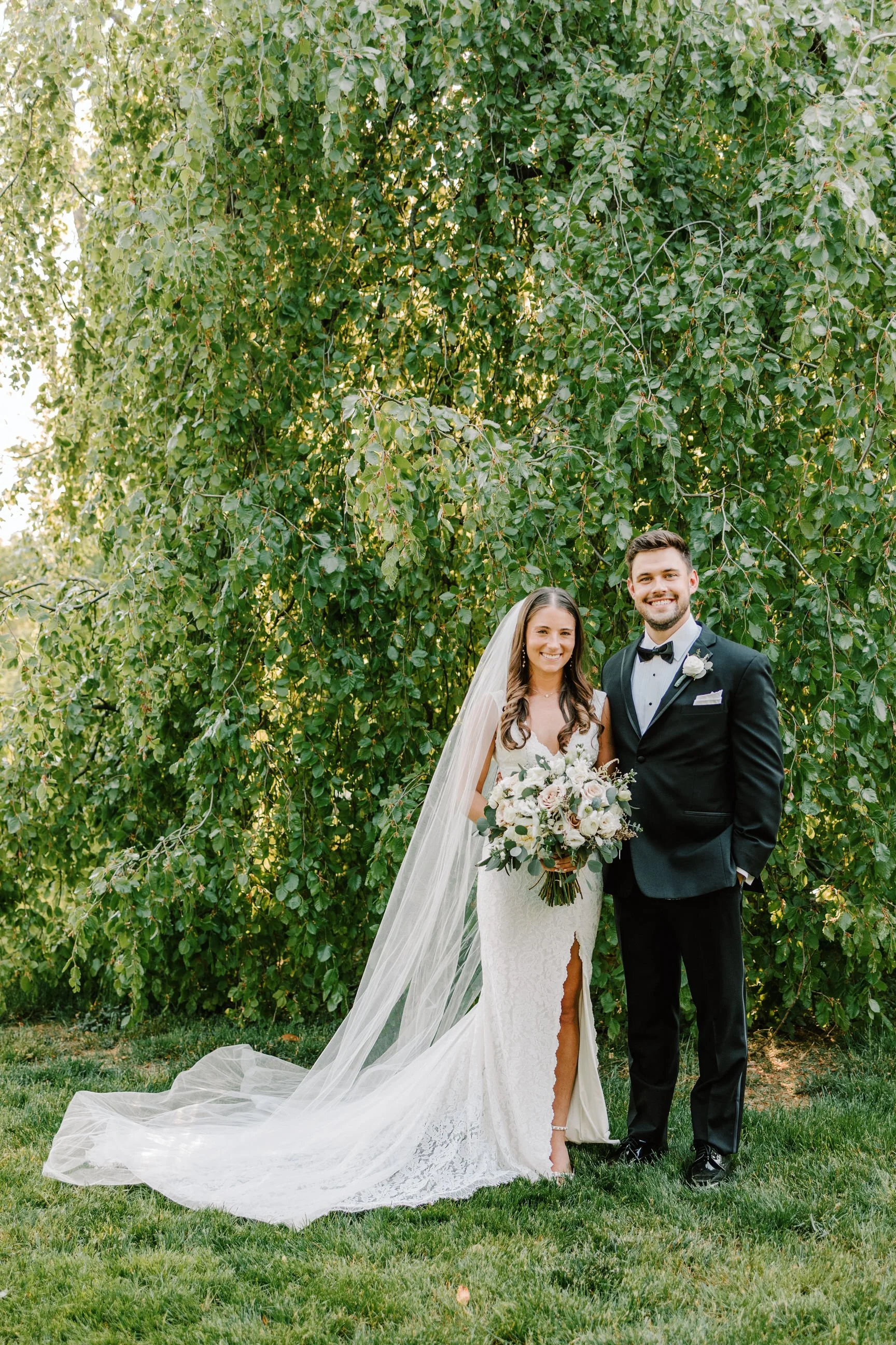 A bride and groom standing outdoors in front of green trees, smiling, dressed in wedding attire with the bride holding a bouquet of flowers.
