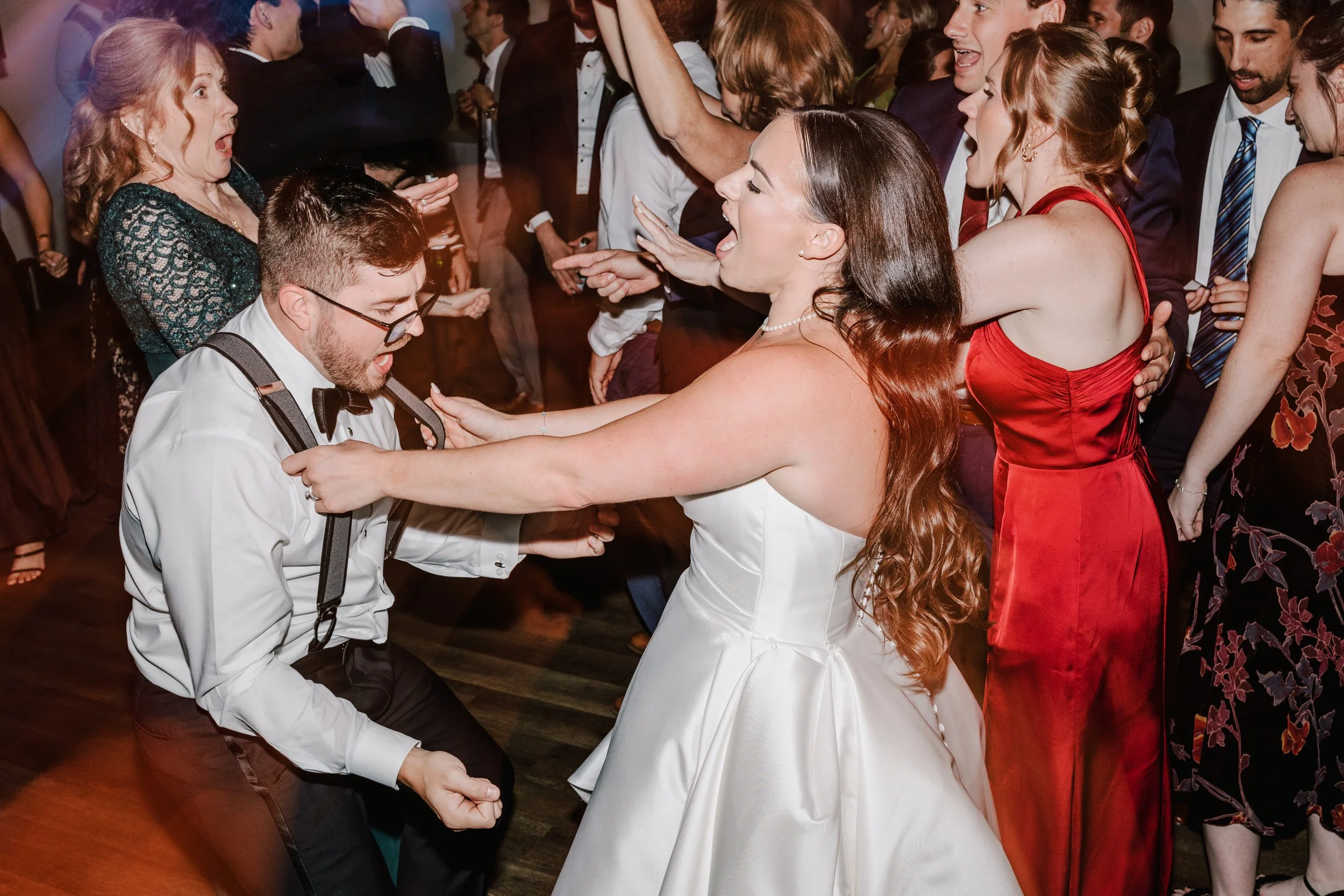 A lively scene of people dancing at a wedding reception, with a woman in a white wedding dress and a man in glasses, suspenders, and a white shirt dancing closely, surrounded by friends in formal attire.