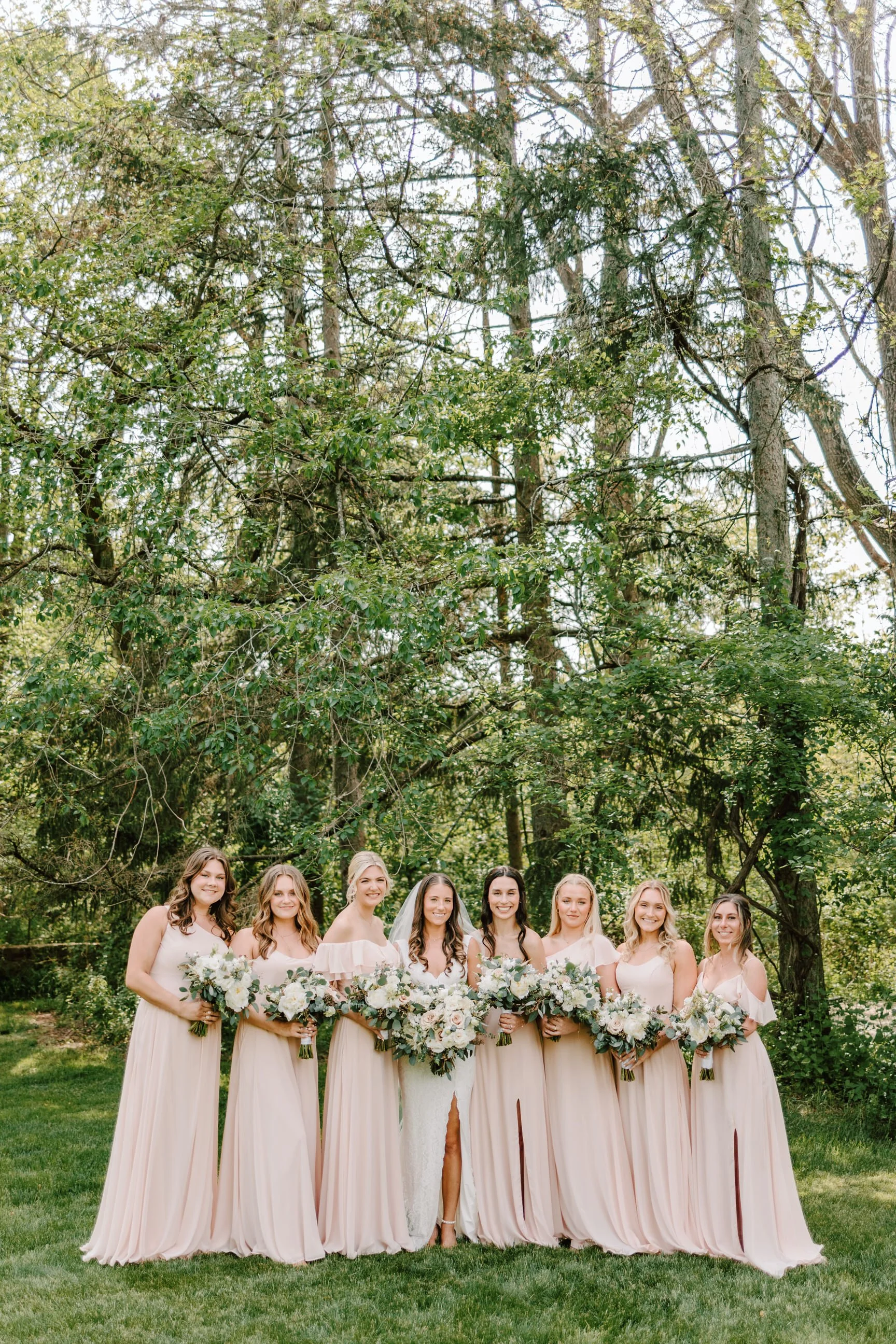Bride and bridesmaids standing outdoors in front of trees, holding bouquets of white and green flowers.