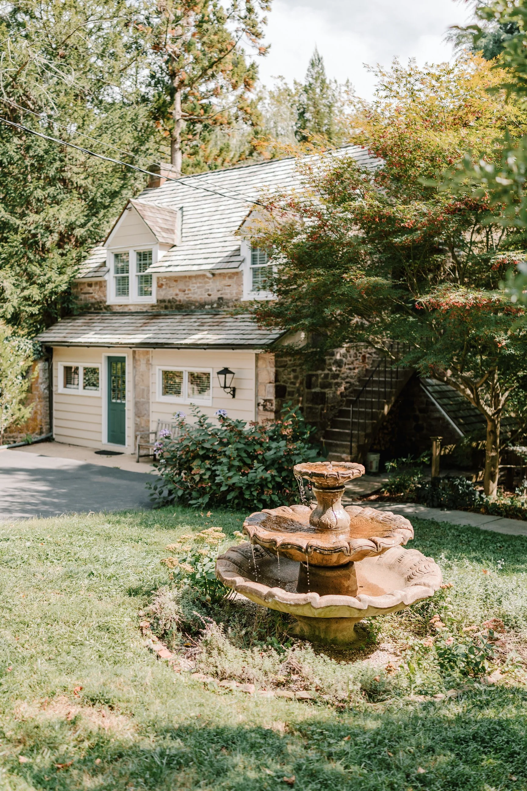 A stone cottage with a slate roof, surrounded by trees, with a fountain in the foreground.