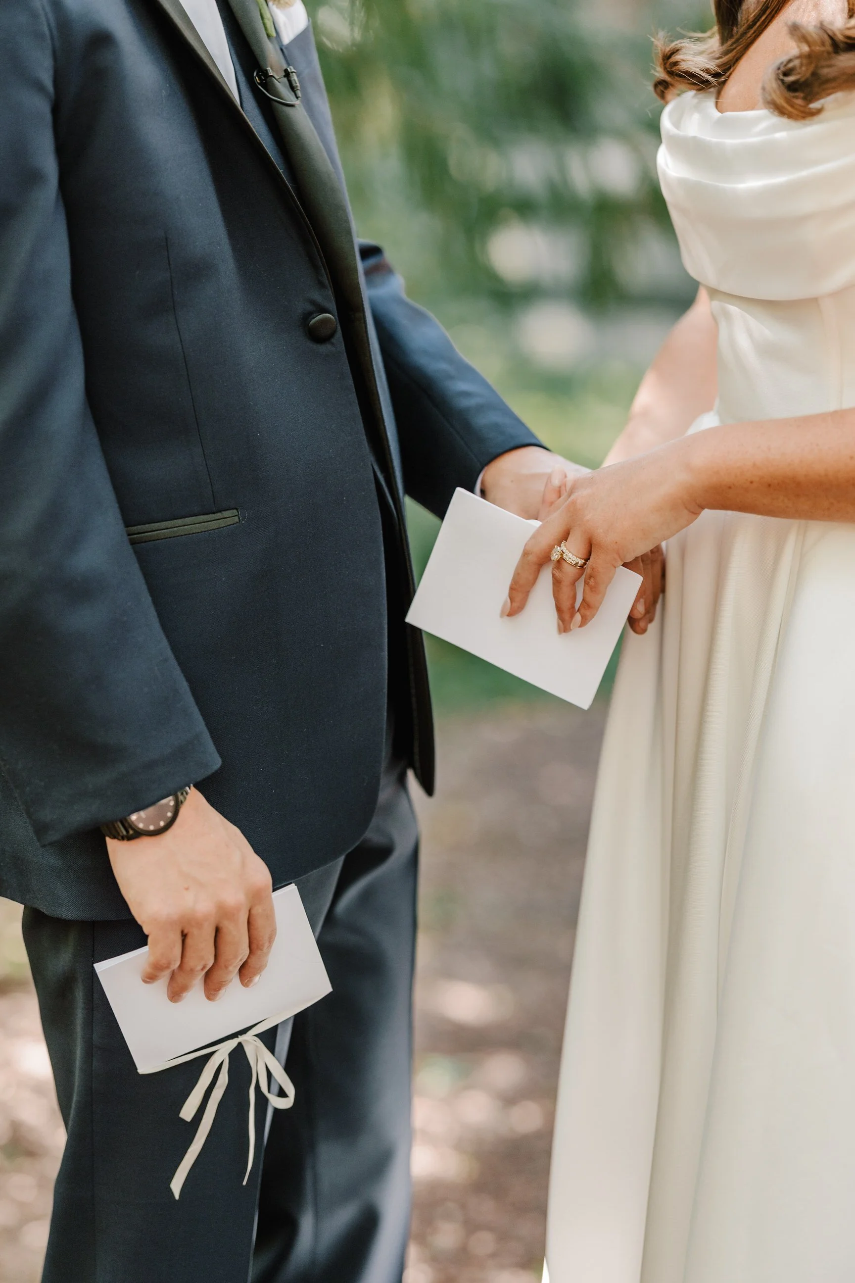 Close-up of a wedding ceremony where a bride and groom are holding hands, holding envelopes. The bride is wearing a white dress and a ring, and the groom is in a dark suit with a dress watch.