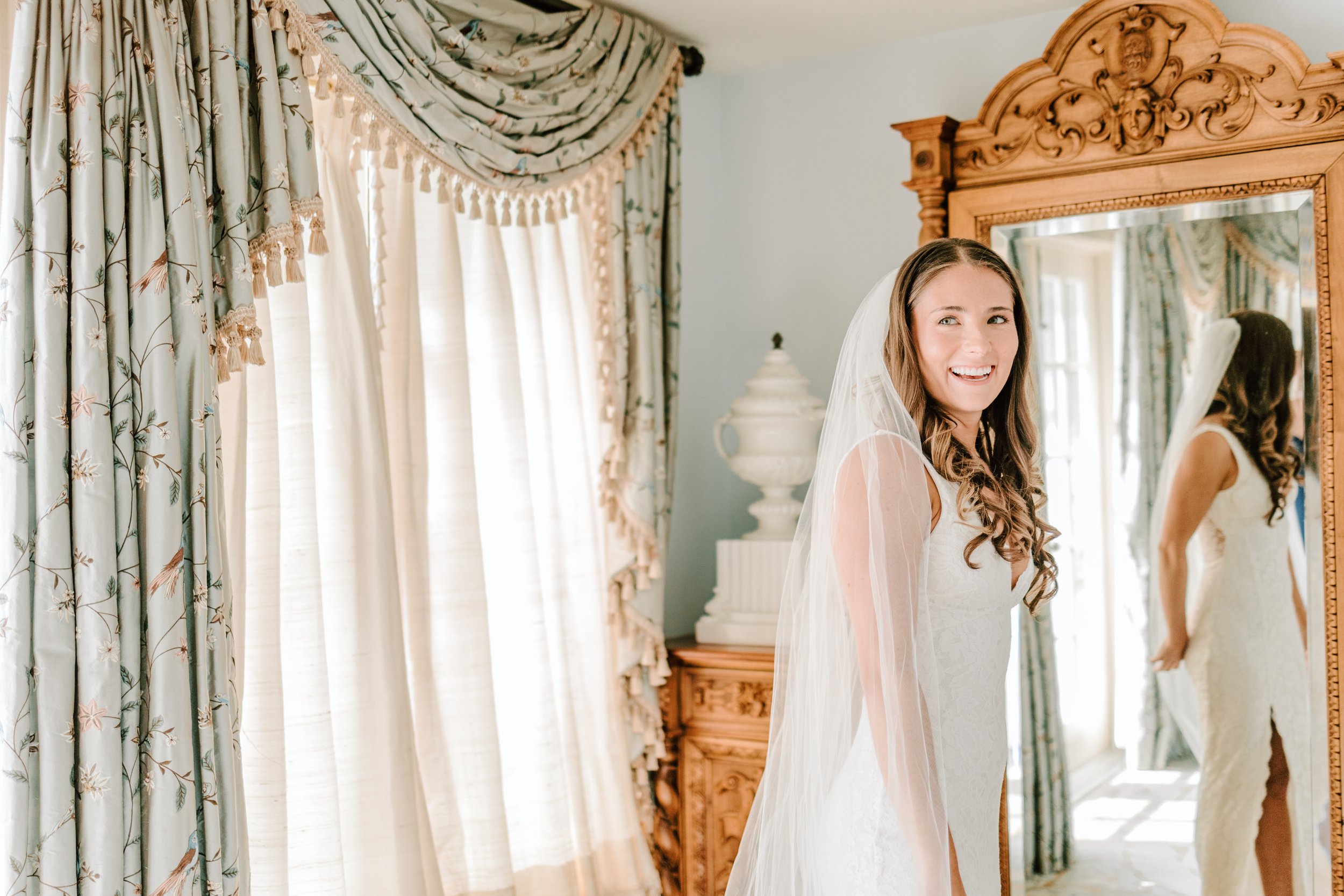 A bride with long, curly hair smiling in a wedding dress and veil, looking into a mirror in a richly decorated room with floral curtains and ornate wooden furniture.