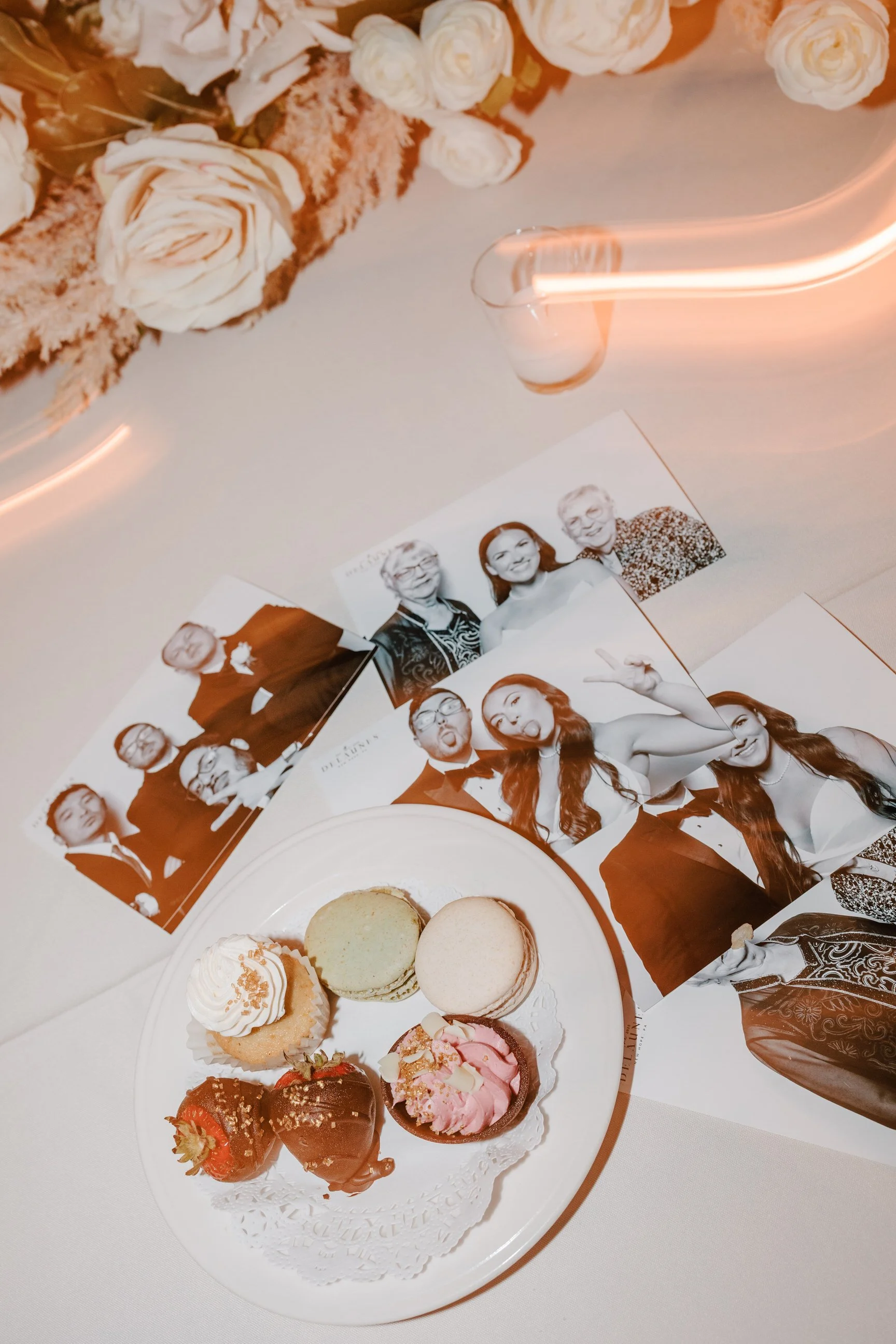 A white plate with six decorated macarons and strawberries, surrounded by black-and-white photo booth pictures of various people, on a decorated table with flowers.