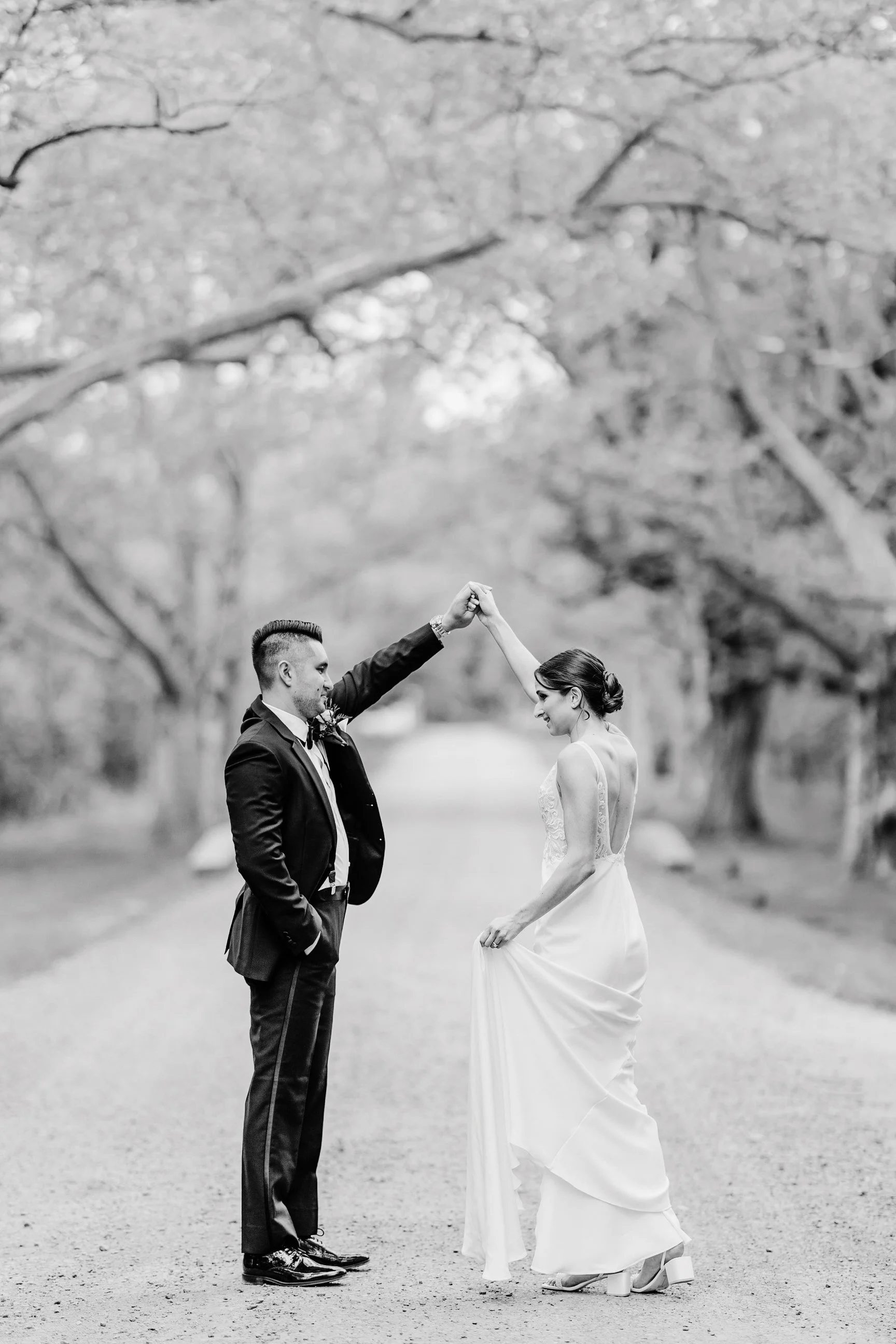 A black and white photo of a bride and groom dancing on a rural path surrounded by trees, with the groom twirling the bride.