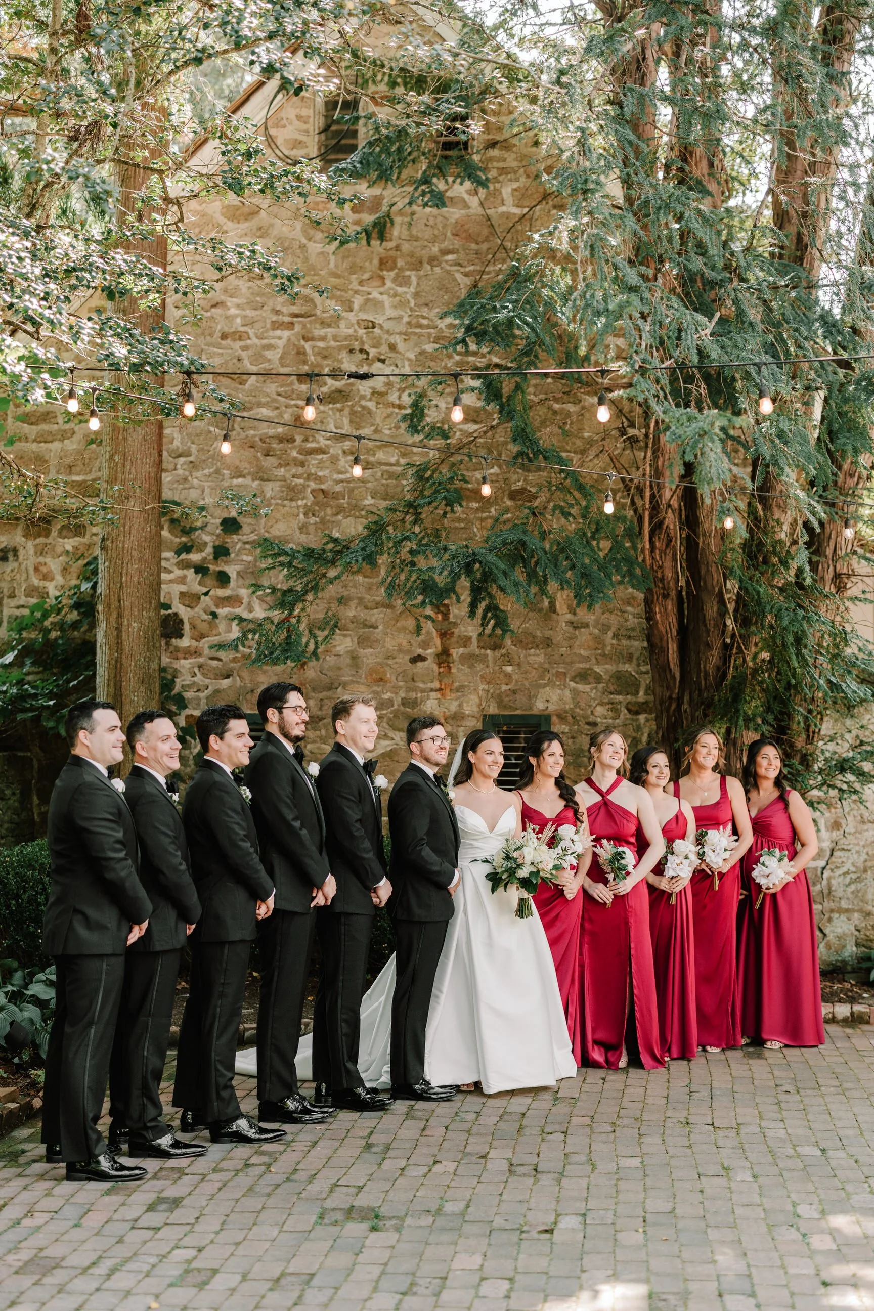 A wedding party of nine people, four men and five women, standing outdoors against a stone wall with trees and string lights overhead. The groom and groomsmen are dressed in black tuxedos, while the bride and bridesmaids are in a white wedding gown a
