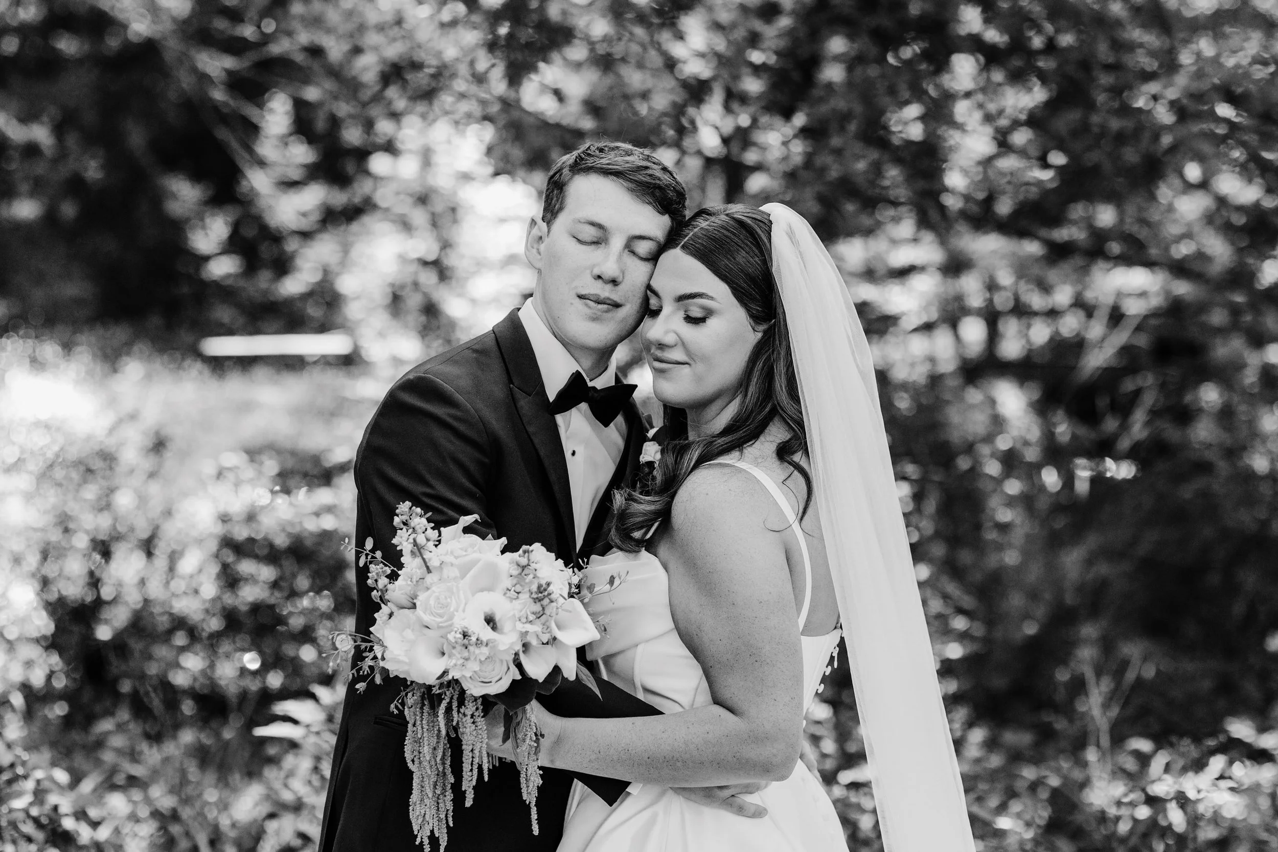 Black and white photo of a bride and groom hugging outdoors, with trees in the background. The groom is wearing a tuxedo with a bow tie, and the bride is holding a bouquet of flowers, wearing a wedding dress and veil.