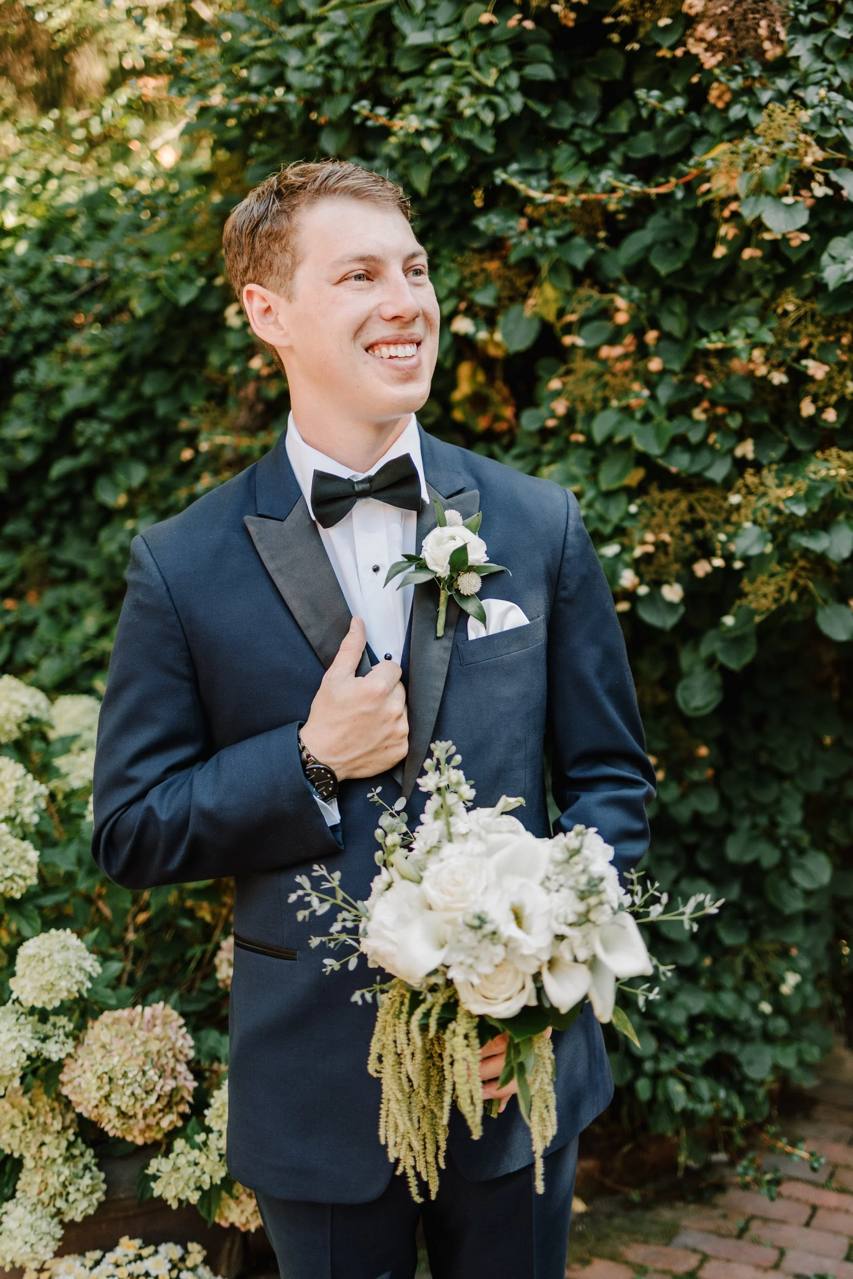 A smiling groom in a navy tuxedo with a black bow tie, holding a bouquet of white flowers, standing outdoors in front of green foliage.